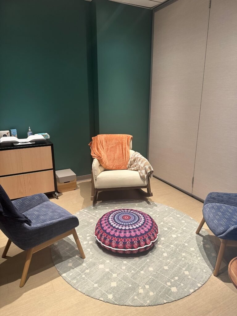A cozy living room corner with two blue chairs, a white armchair with an orange towel, a patterned round cushion on a gray area rug, and a wooden cabinet against a green wall.