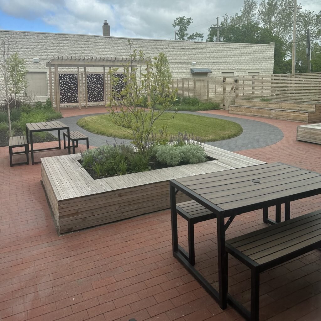 A backyard patio with wooden benches, tables, a small grassy area, some plants, and a wooden fence, under a partly cloudy sky.