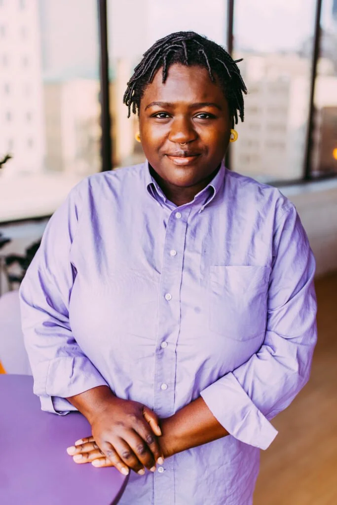 A woman with short dreadlocks and yellow earrings wearing a light purple shirt, standing indoors near large windows with a cityscape view.