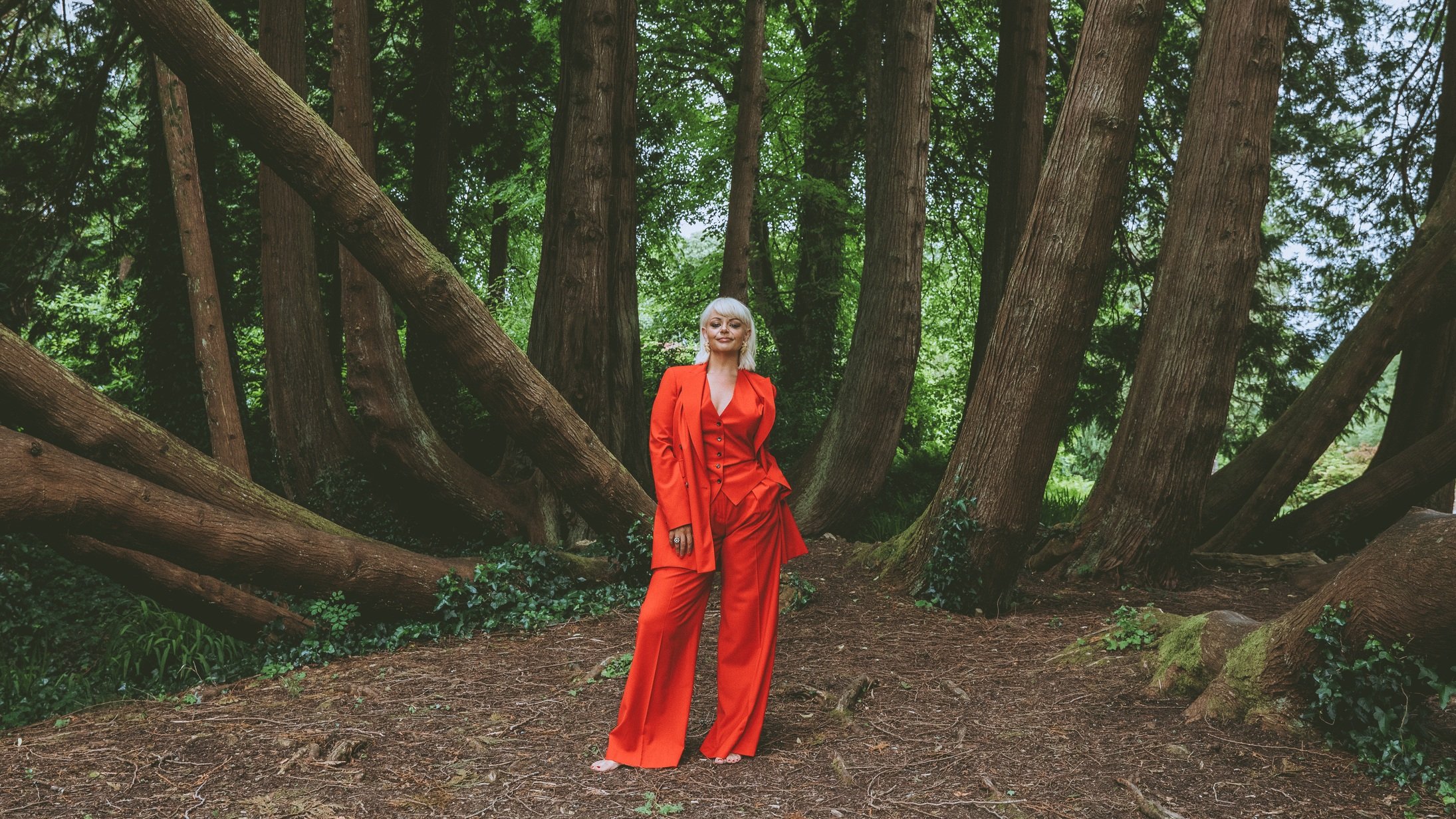 A woman in a bright orange suit standing in a lush green forest with large trees and dense foliage.