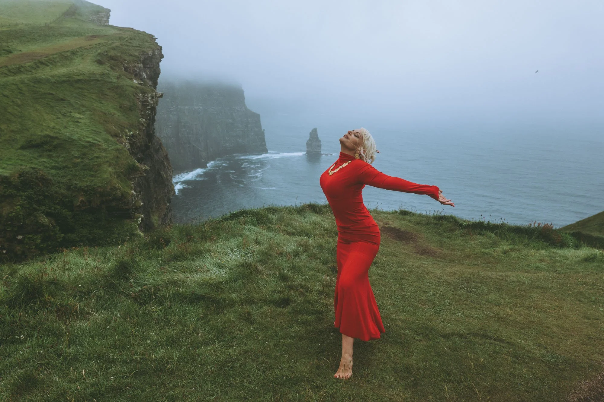 A woman in a red dress standing on a grassy hill with arms outstretched, facing the ocean and cliffs shrouded in fog.