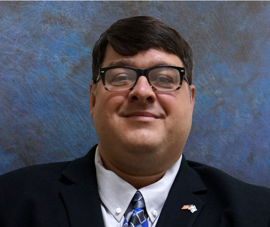 A man in a business suit, wearing glasses and a tie, smiling at the camera, with an American flag lapel pin, against a mottled blue background.