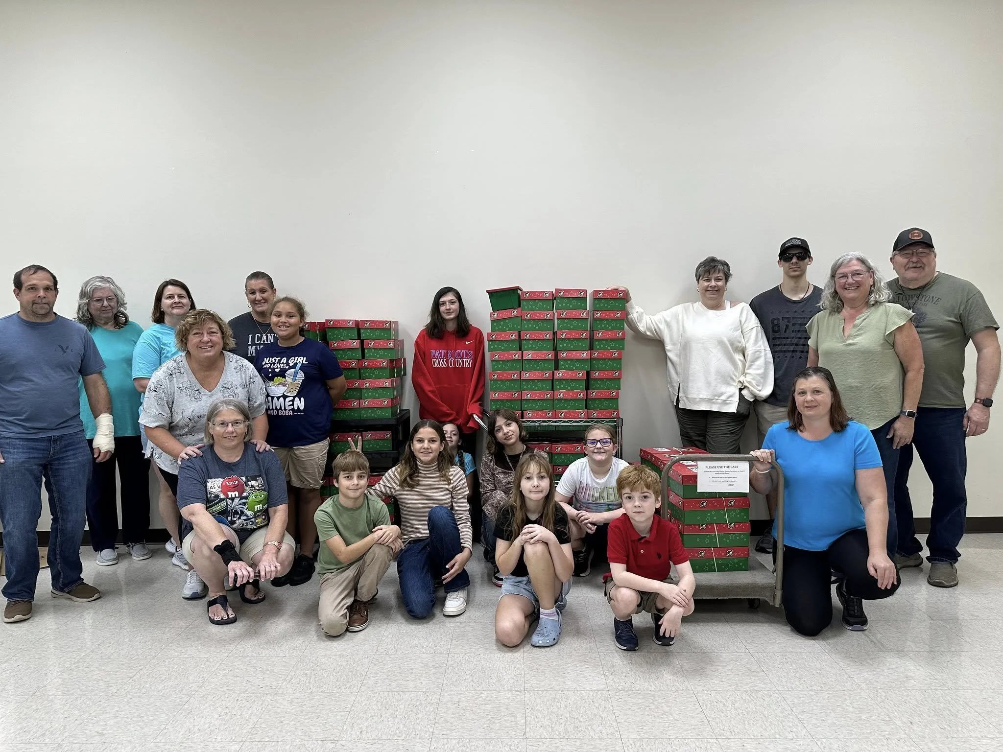 A group of people, including adults and children, gathered together in front of stacks of Christmas-themed boxes and a cart, for a holiday charity event or celebration.