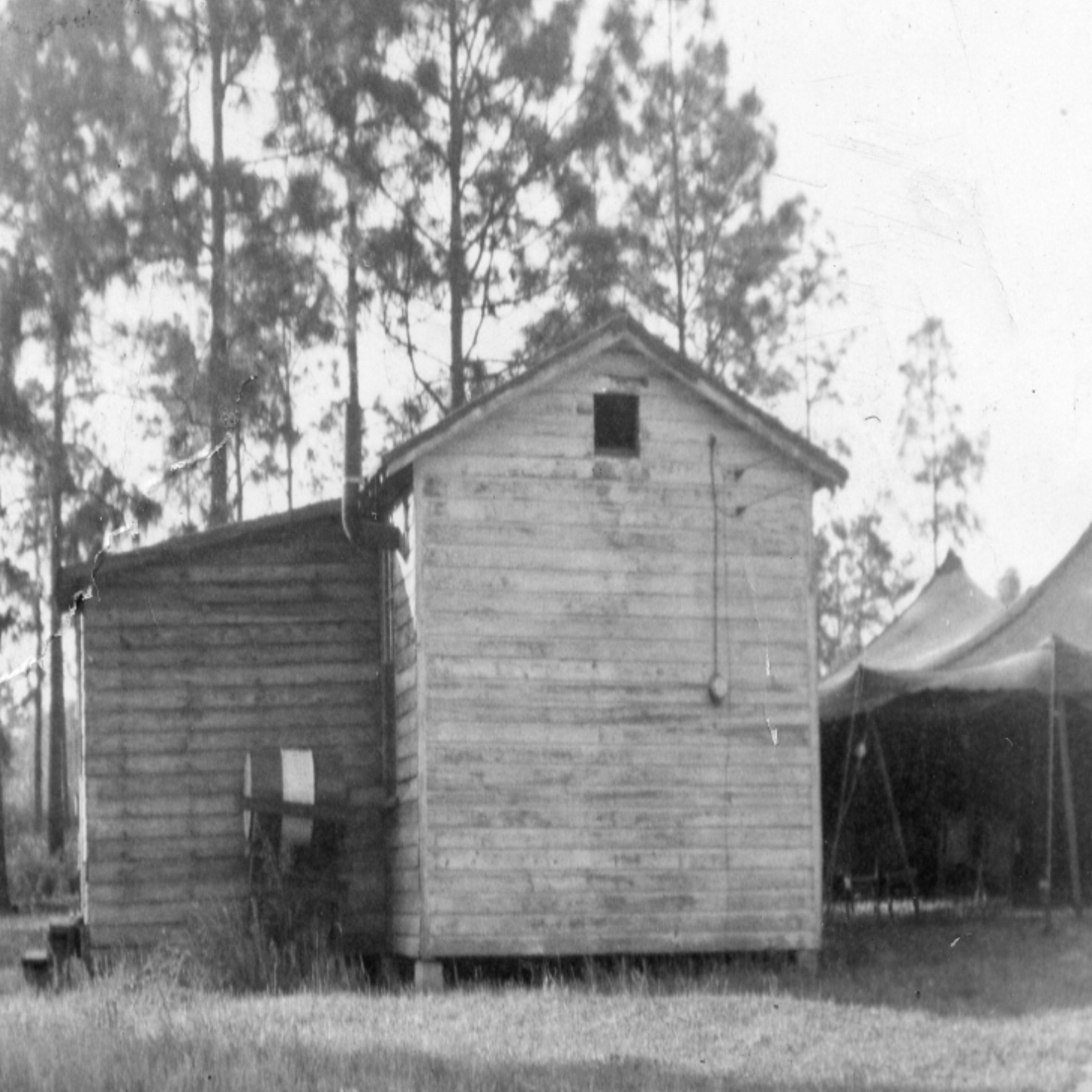 An old black and white photo of a small wooden building, possibly a barn or shed, with a pitched roof and a small rectangular opening near the top. There are tall trees in the background and a tent-like structure to the right.