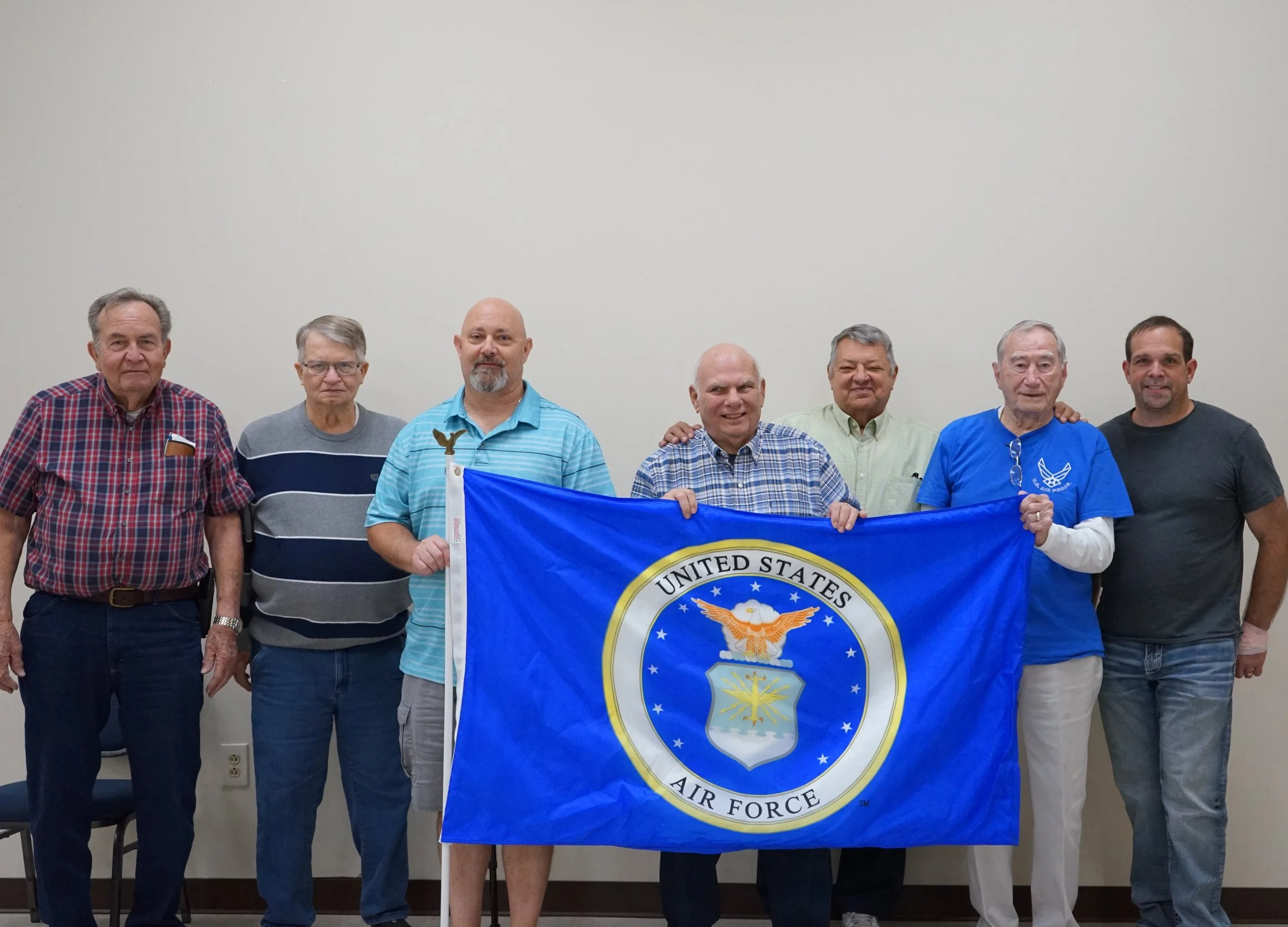 A group of seven men standing indoors against a plain white wall, holding a blue United States Air Force flag. The men are dressed casually, with some wearing plaid or striped shirts and one in a blue T-shirt with a logo. One man in the center is smi
