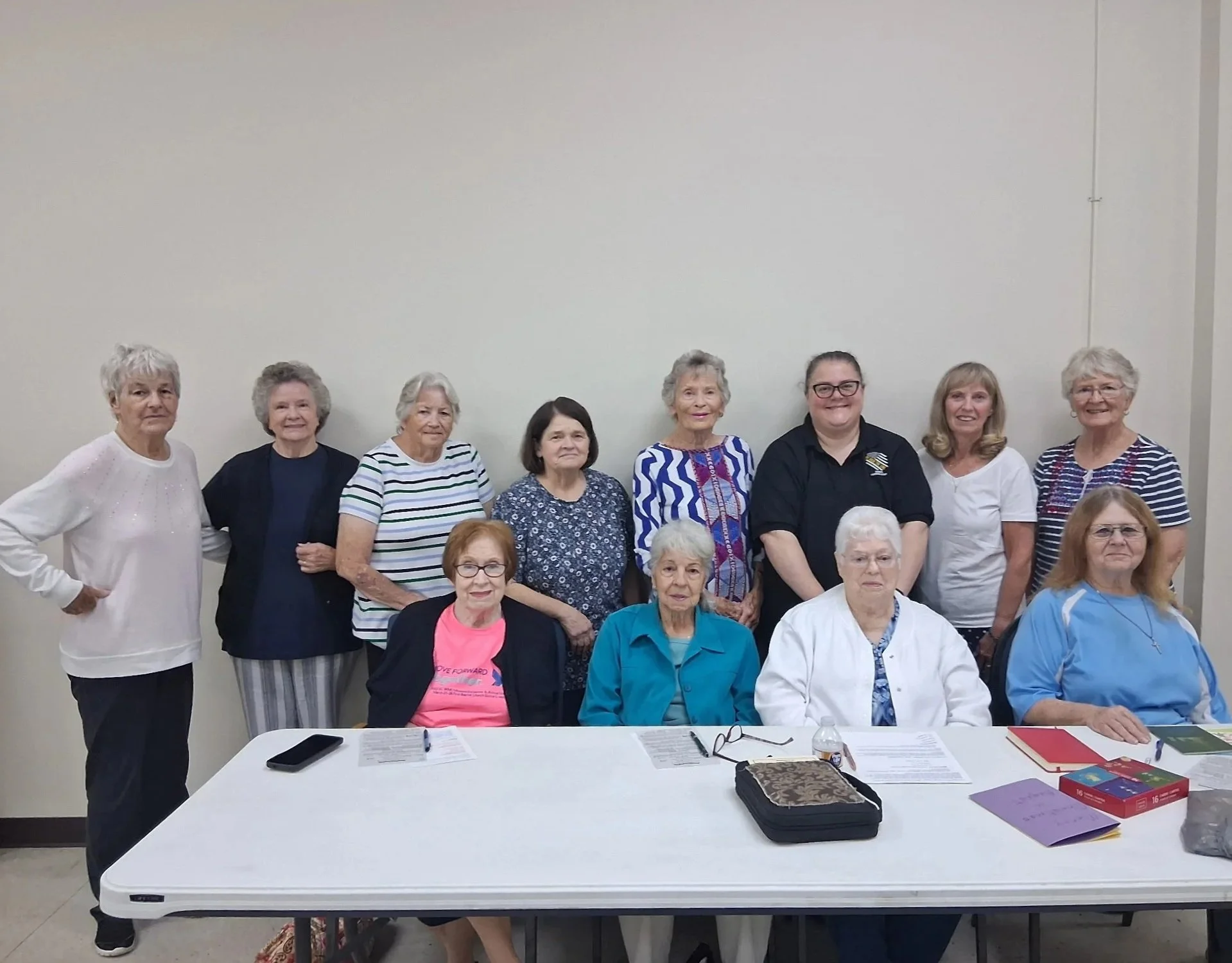 Group of thirteen women, most elderly, posing for a photo in a meeting room with a white wall background, some seated at a table with documents and books, others standing behind.