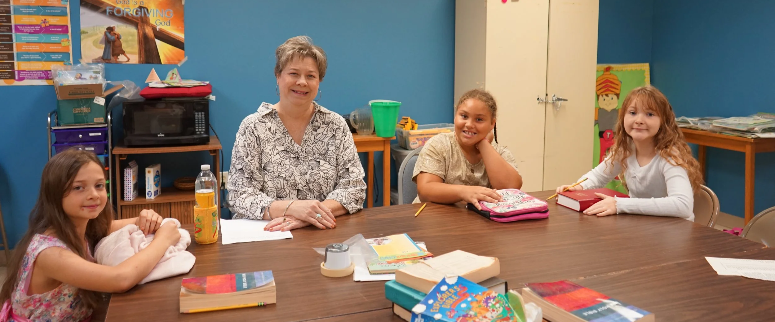 A classroom scene with four female students and a female teacher sitting at a large table. The students are smiling, and the classroom has educational posters, books, and supplies visible.