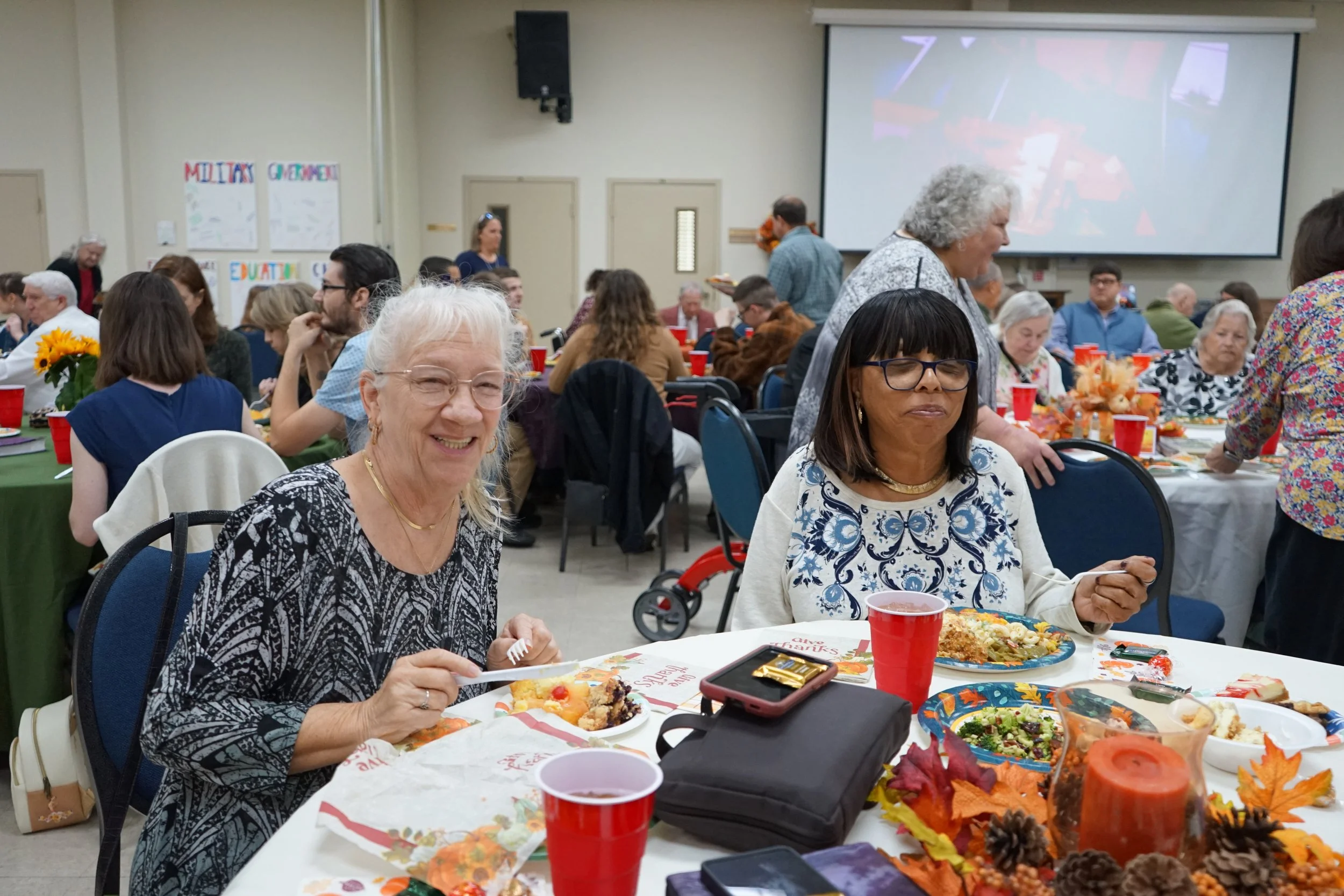 Two women sitting at a table during a large social gathering or celebration. They are eating food, with plates of meal and red cups in front of them. The woman on the left is smiling, wearing glasses and a patterned black and white top, while the wom