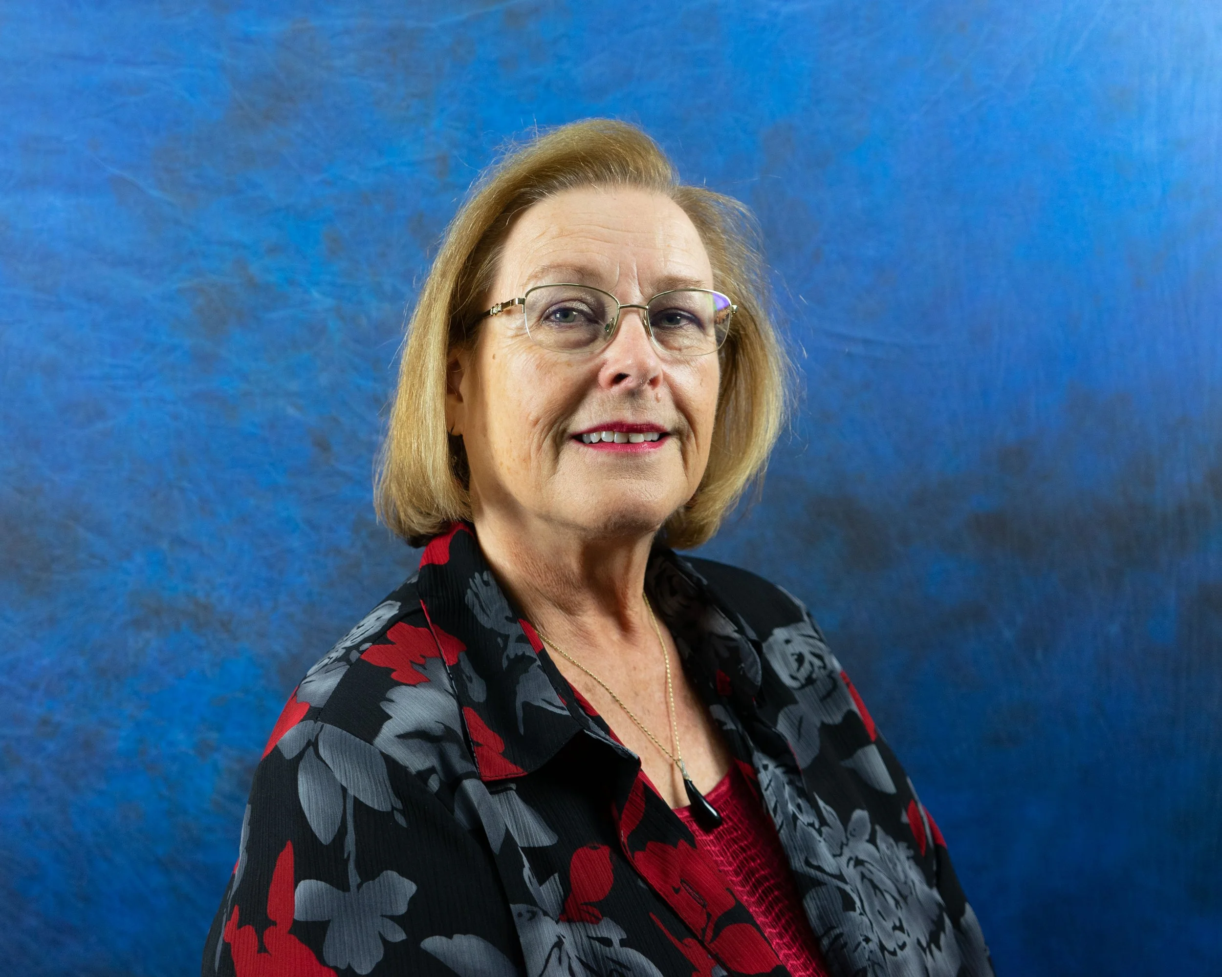 A woman with glasses and shoulder-length blonde hair smiling at the camera, wearing a black and red floral blazer over a red top, standing against a blue textured background.