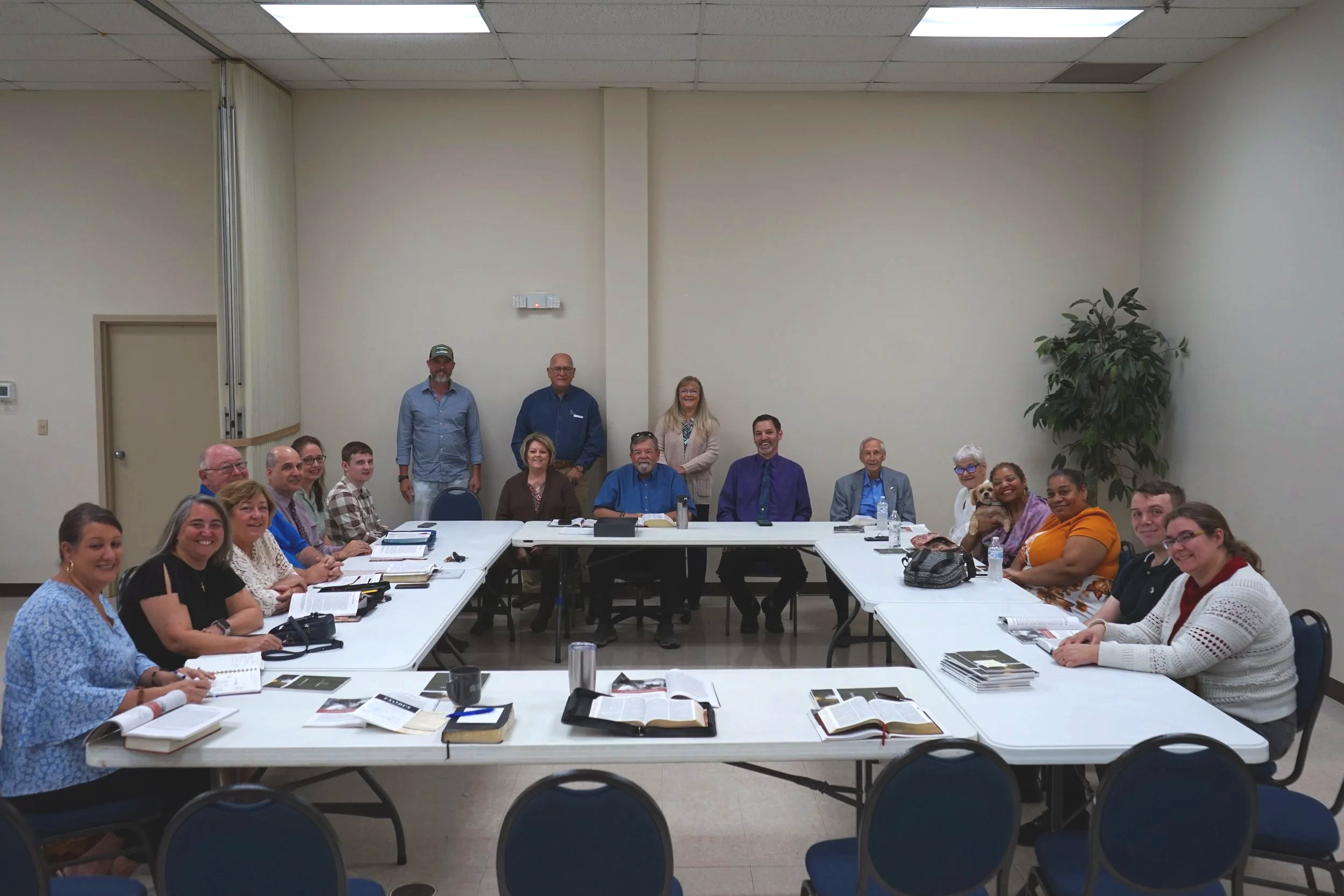 People seated around a large rectangular conference table in a meeting room with a few standing in the back.