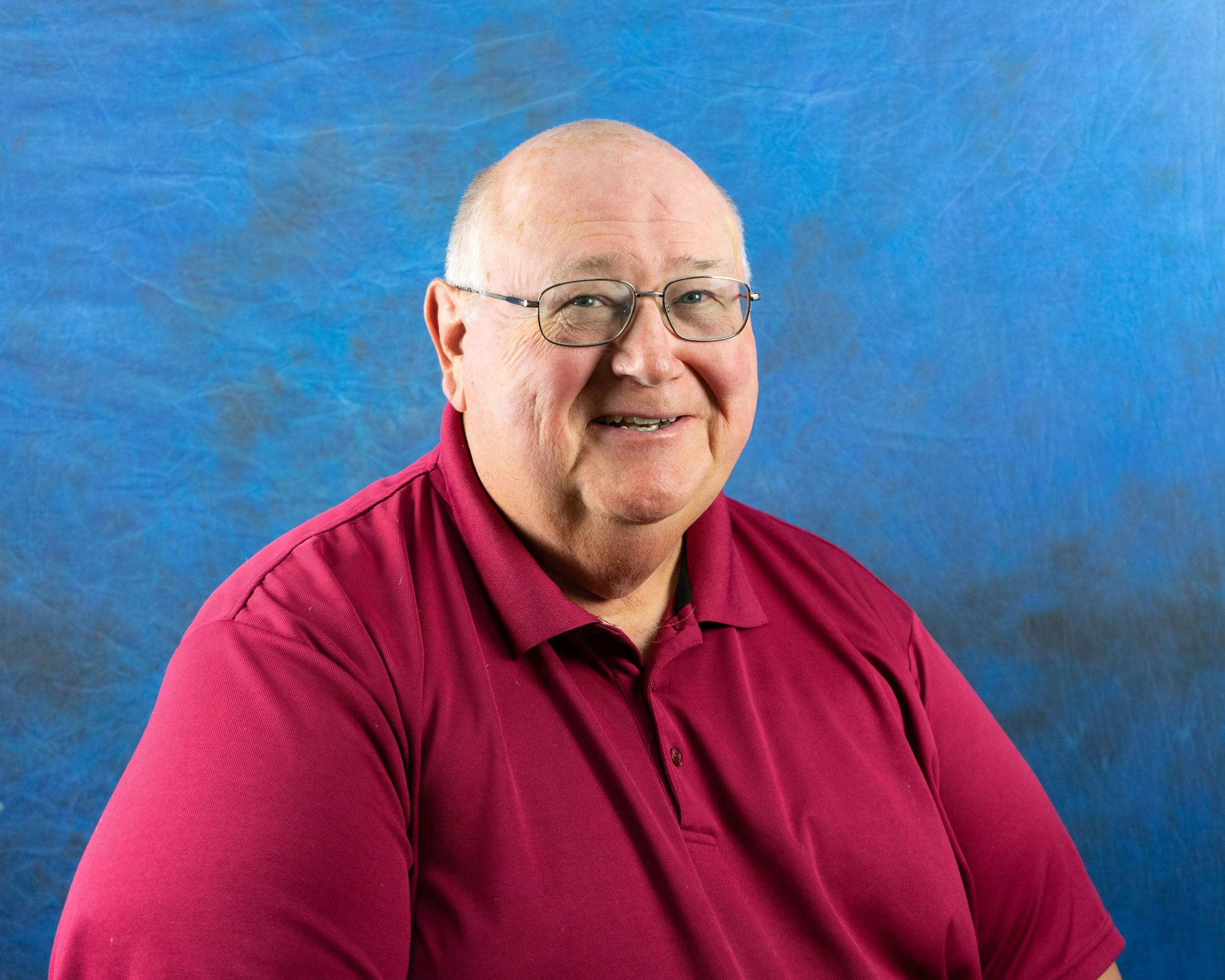 A smiling man with glasses and a maroon polo shirt sitting against a blue background.