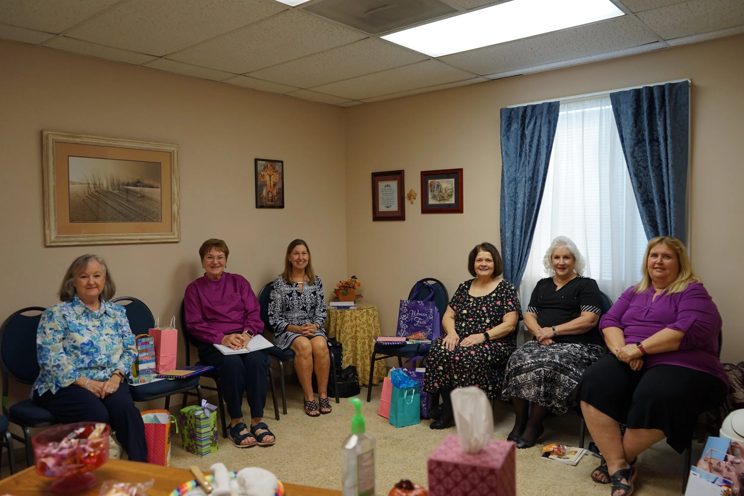 Six women sitting in a circle in a room with beige walls, blue curtains, and framed pictures on the wall, smiling and attending a gathering with gift bags and presents around them.