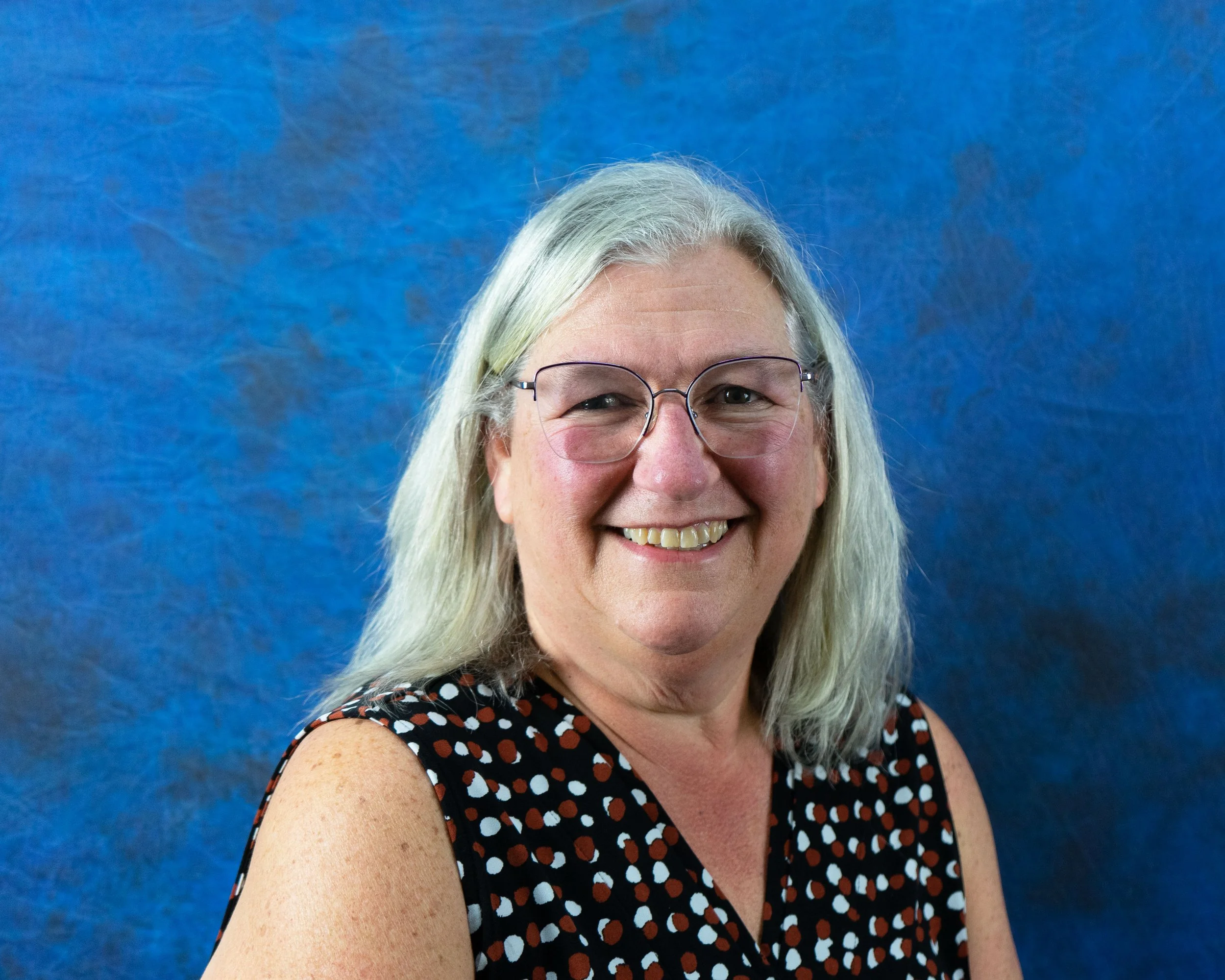 A smiling middle-aged woman with glasses and shoulder-length gray hair, wearing a sleeveless black top with a red and white polka dot pattern, against a blue textured background.