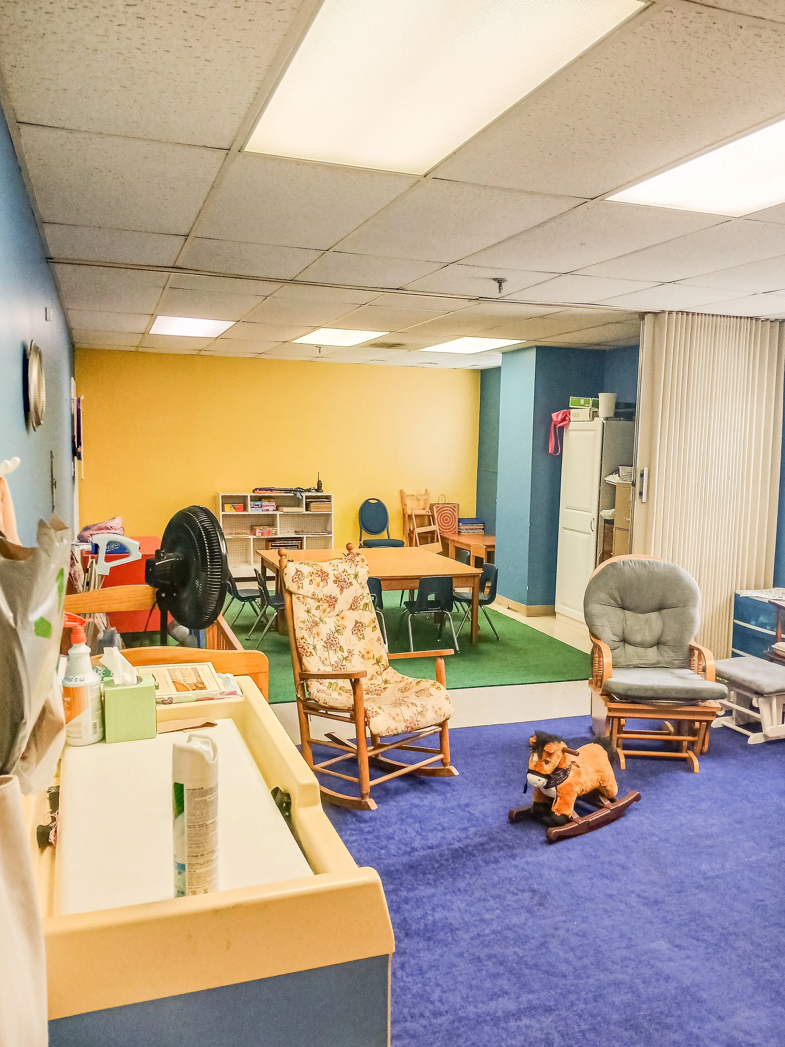 A cozy room with colorful walls, including yellow, blue, and green sections. The room has various chairs, a table, and a plush rocking horse. It appears to be a reading or activity room, with a calendar and supplies visible on the shelves. The ceiling has rectangular lights, and there is a small standing fan in the room.
