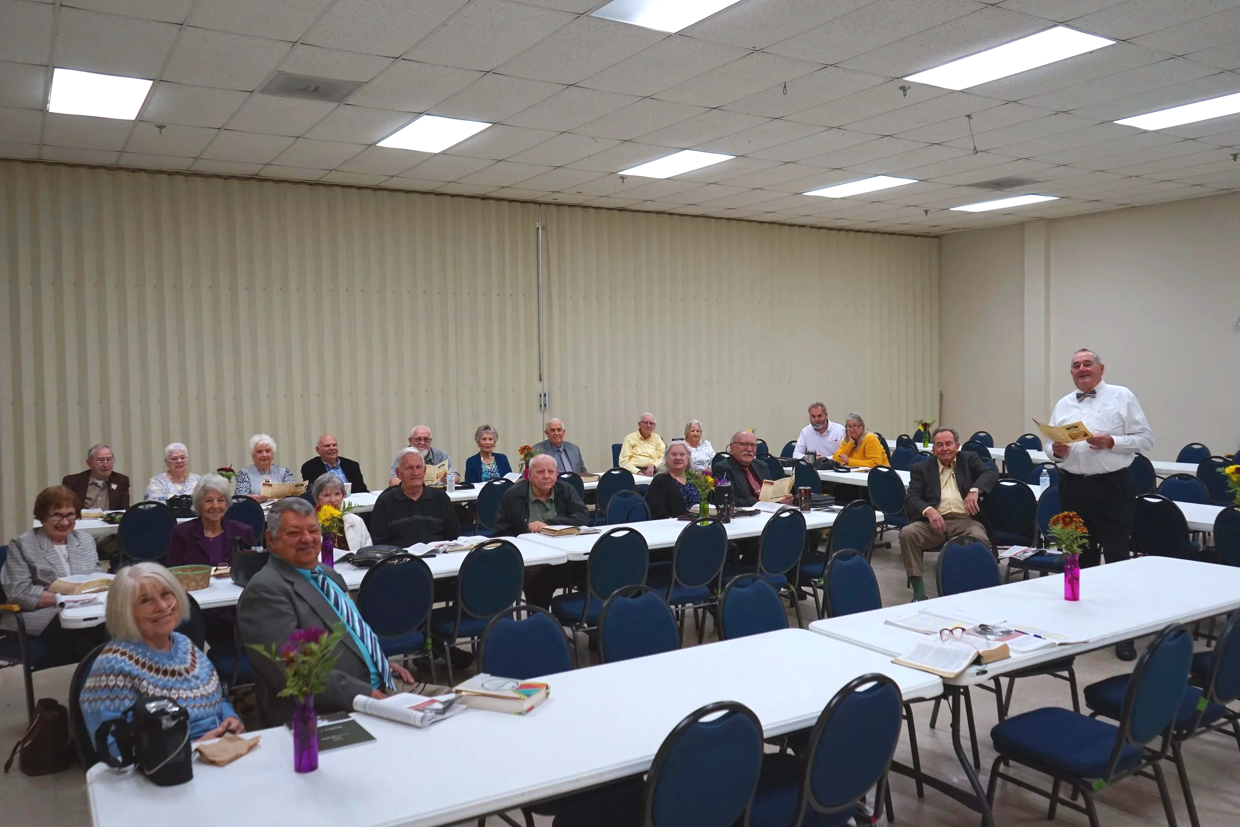 A group of elderly people seated at long tables in a banquet hall, listening to a man in a white shirt and bow tie speaking at the front.