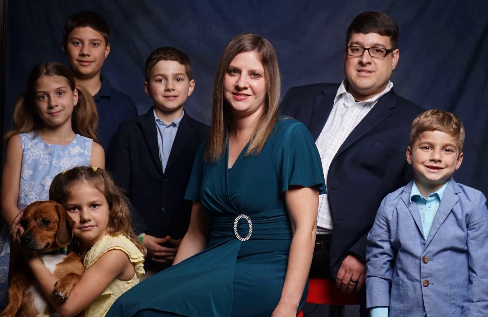 Group portrait of a family with six children and a dog, all dressed in formal attire, standing against a dark backdrop.