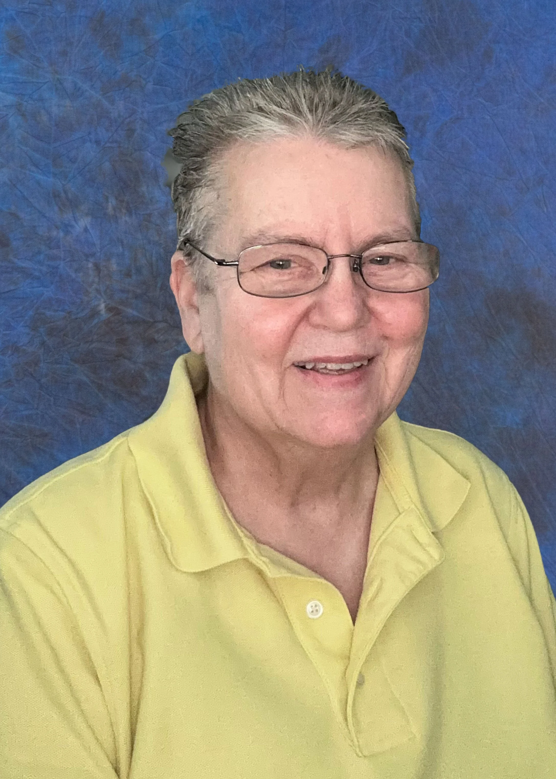 A smiling older woman with short gray hair, glasses, wearing a yellow collared shirt, against a textured blue background.