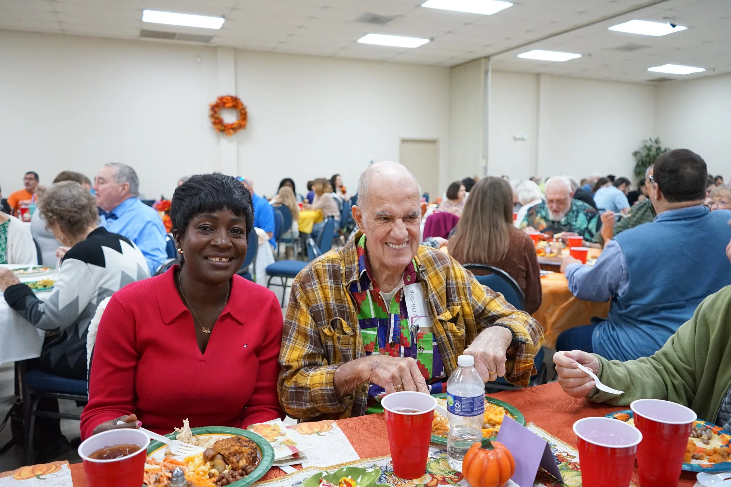 Two people, a woman in a red shirt and a man in a plaid shirt, sitting at a table during a festive gathering, enjoying a meal in a large decorated banquet hall filled with many other attendees.