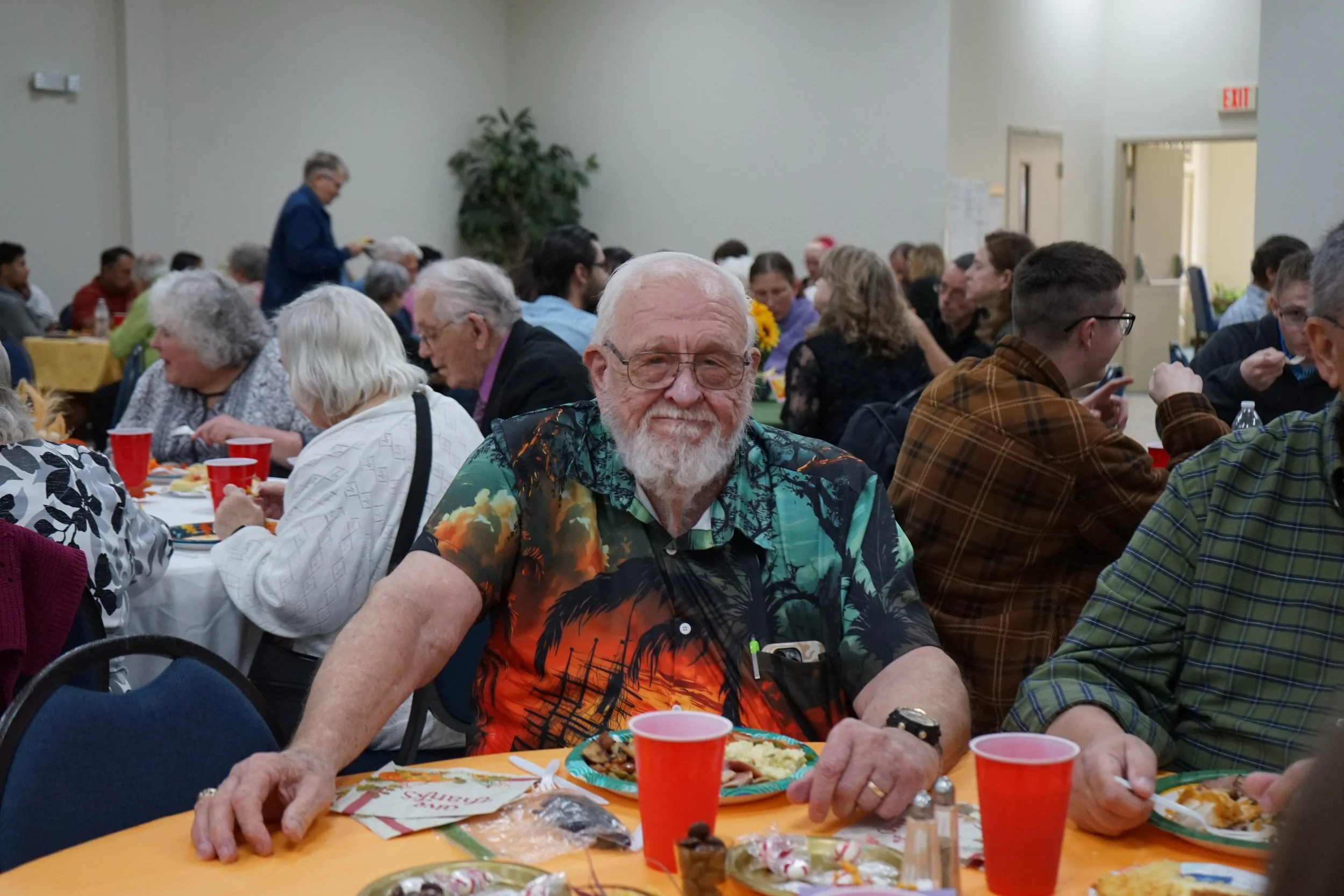 An elderly man with a beard and glasses sitting at a table with food and drinks during a gathering or celebration in a large hall filled with people.