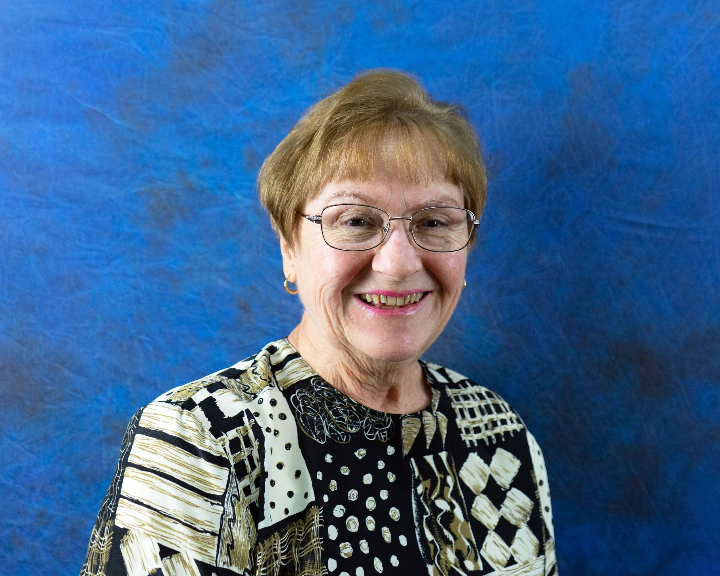 Senior woman with glasses smiling, wearing a patterned black and gold top, against a textured blue background.
