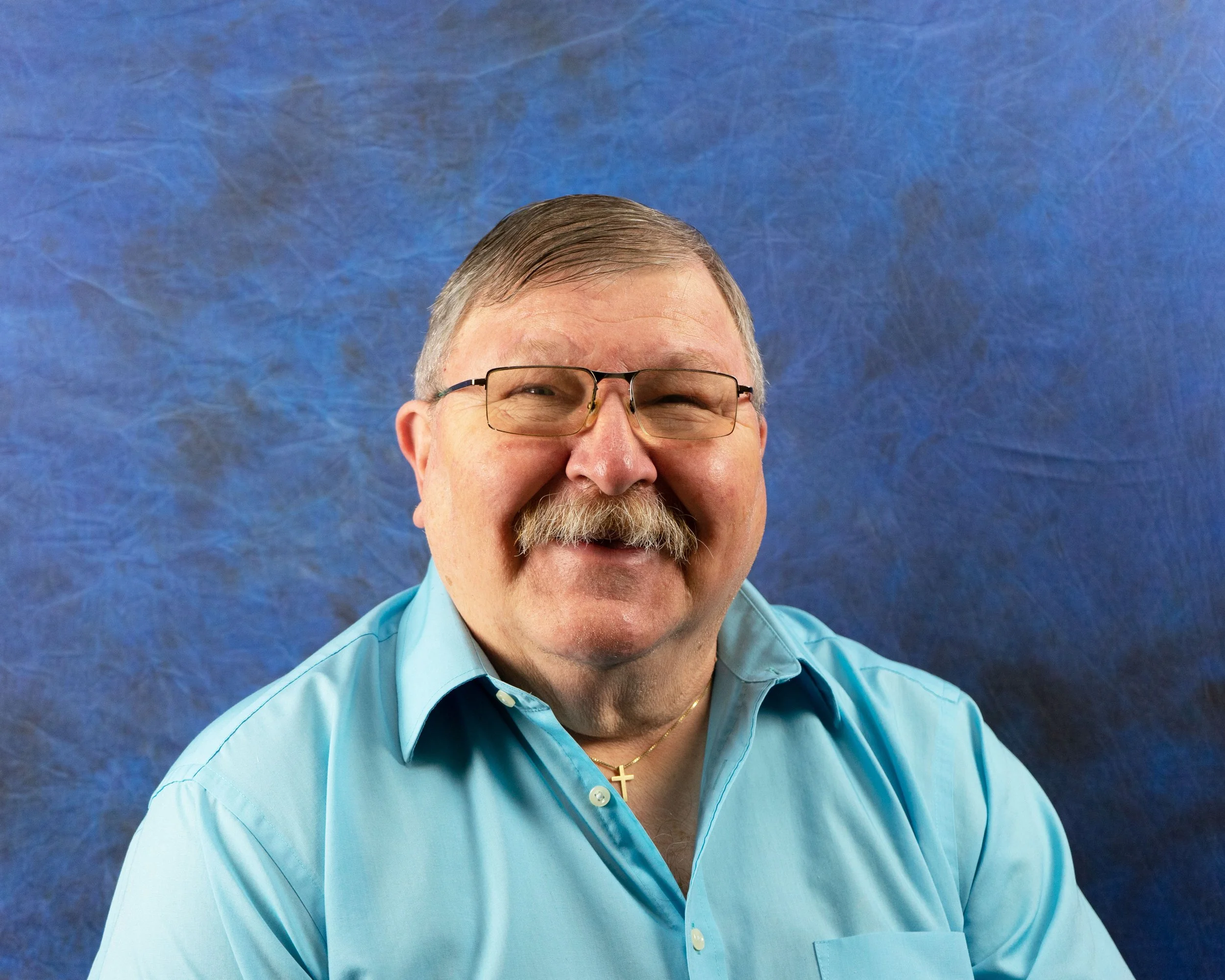A smiling middle-aged man with gray hair, mustache, glasses, wearing a light blue button-up shirt, and a gold cross necklace, against a textured blue background.