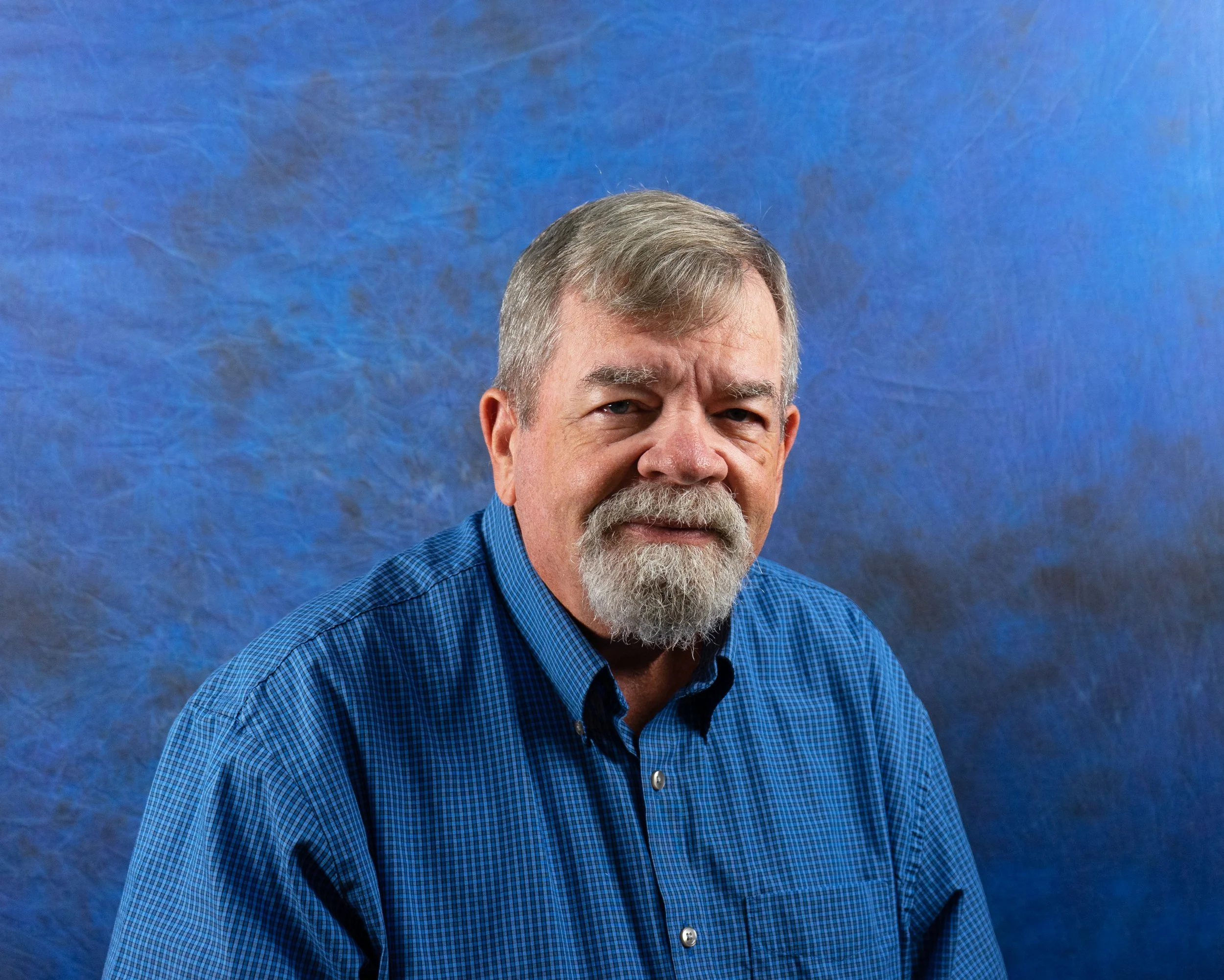 A middle-aged man with gray hair, beard, and mustache wearing a blue checkered shirt poses against a textured blue background.