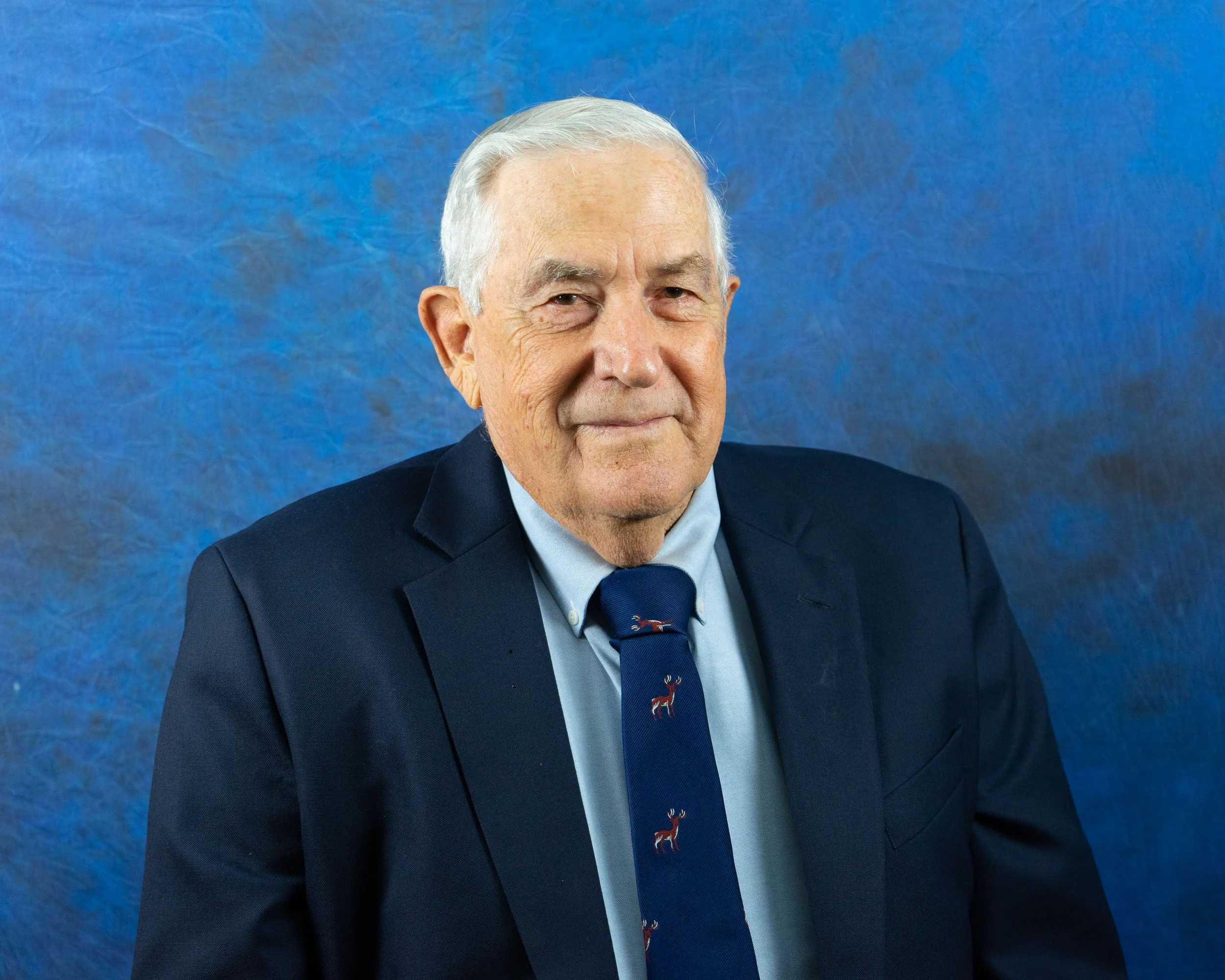 Older man with gray hair smiling, dressed in a dark blue suit, light blue shirt, and a blue tie with red reindeer, standing against a blue textured background.