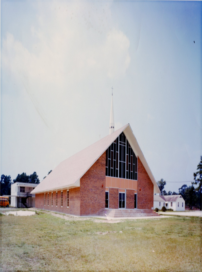 A church with a brick exterior, large stained glass windows, and a tall white steeple with a cross, situated on a grassy lawn.