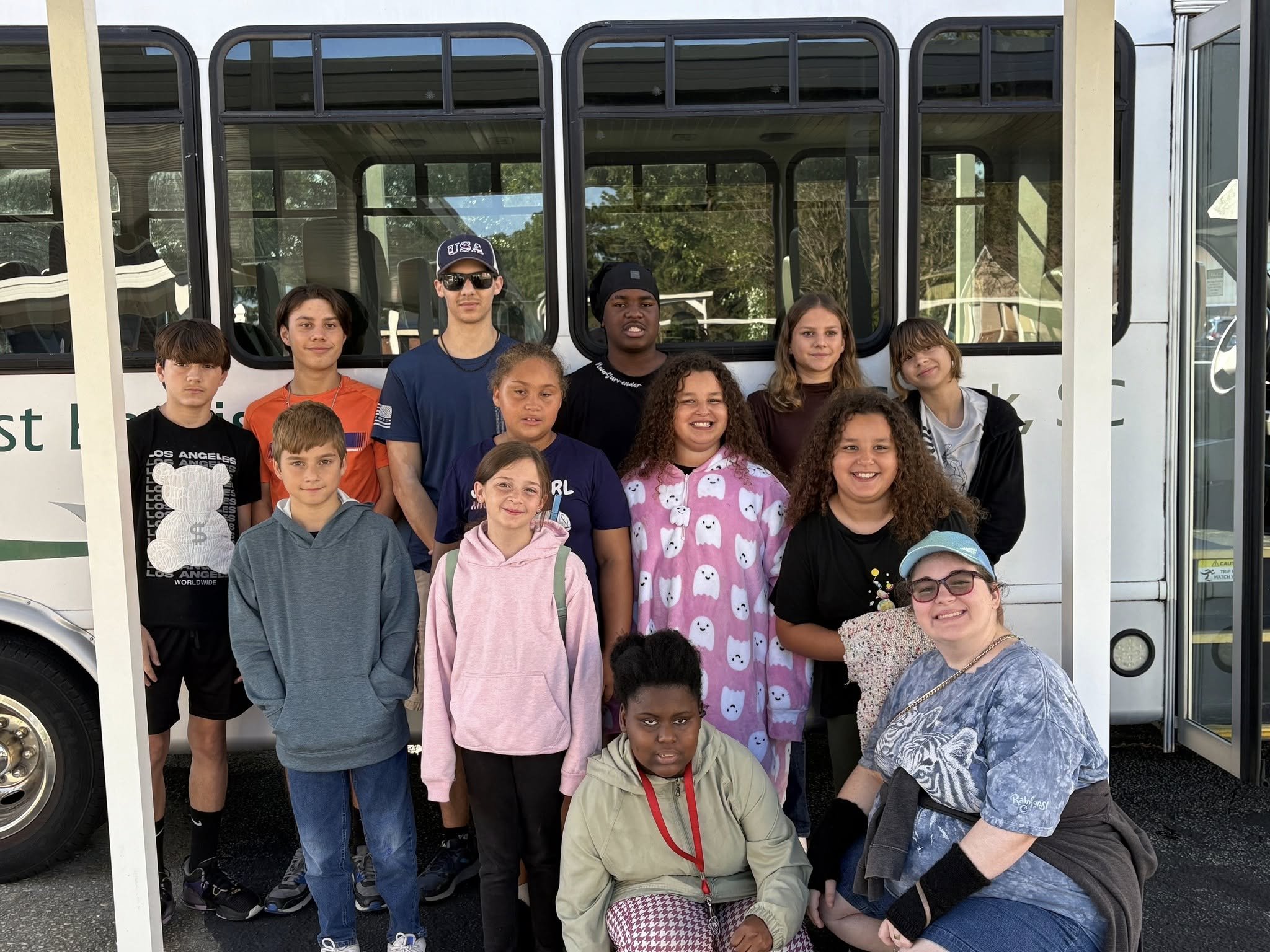 Group of children and teenagers posing in front of a bus, some smiling and some with neutral expressions, with a woman kneeling in front, wearing glasses and a blue cap.