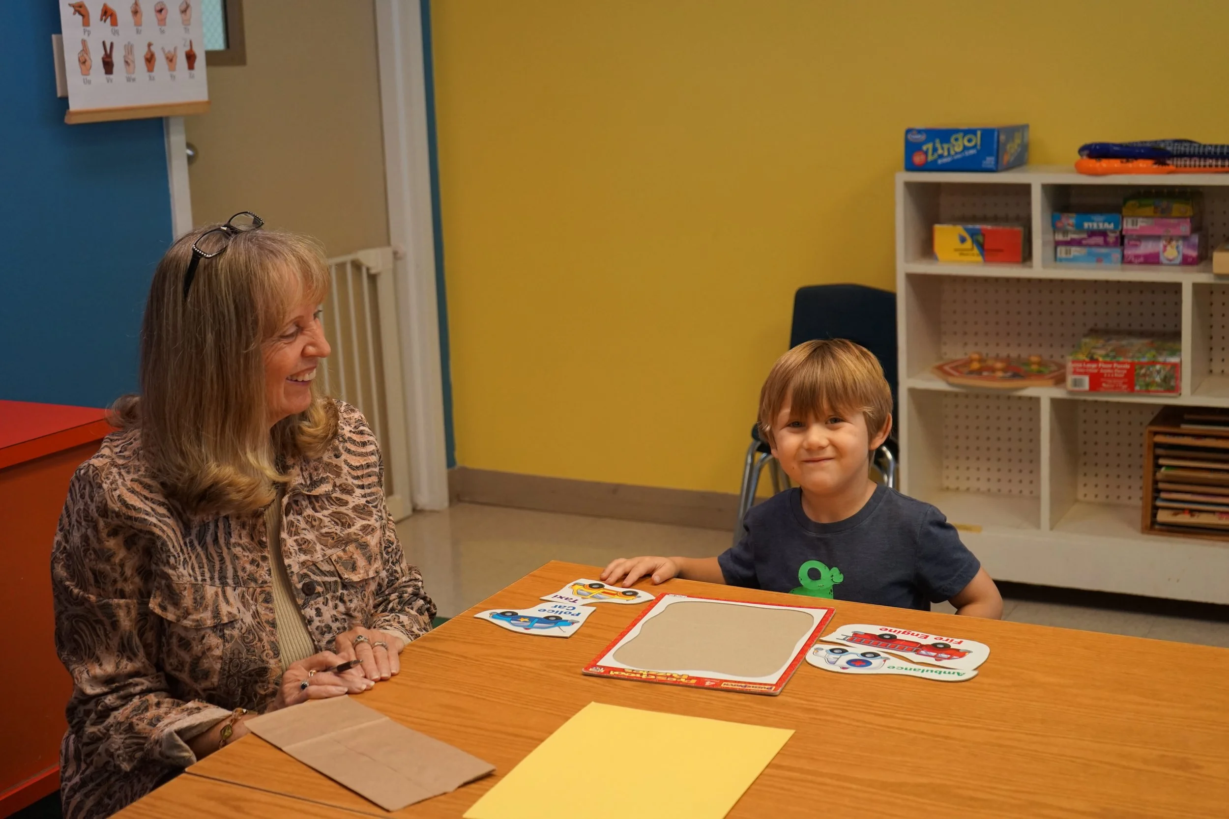 A woman and a young boy sitting at a table in a classroom, smiling. The table has colorful puzzle pieces and a yellow piece of paper. Shelves with games and toys are in the background.