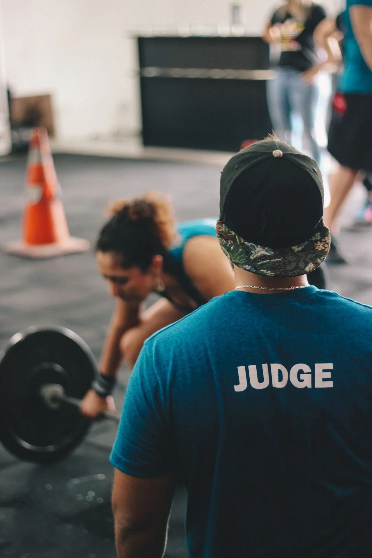 A man with a black baseball cap and a blue shirt with the word JUDGE on the back, standing in a gym watching a woman doing a deadlift exercise.