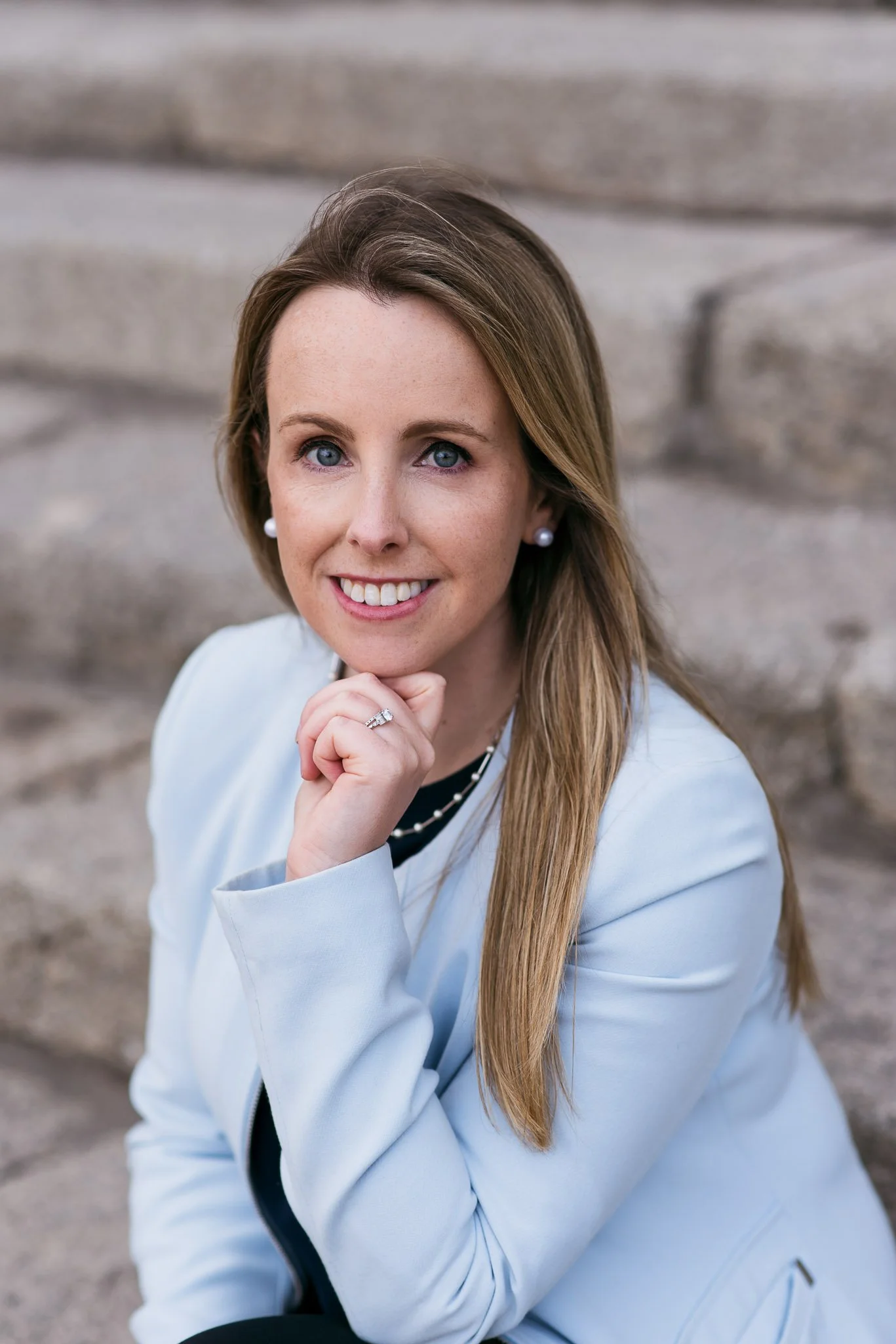 A portrait of a woman with long blonde hair, blue eyes, and a smile, wearing pearl earrings, a ring, and a light blue blazer, sitting outdoors with stone steps in the background.