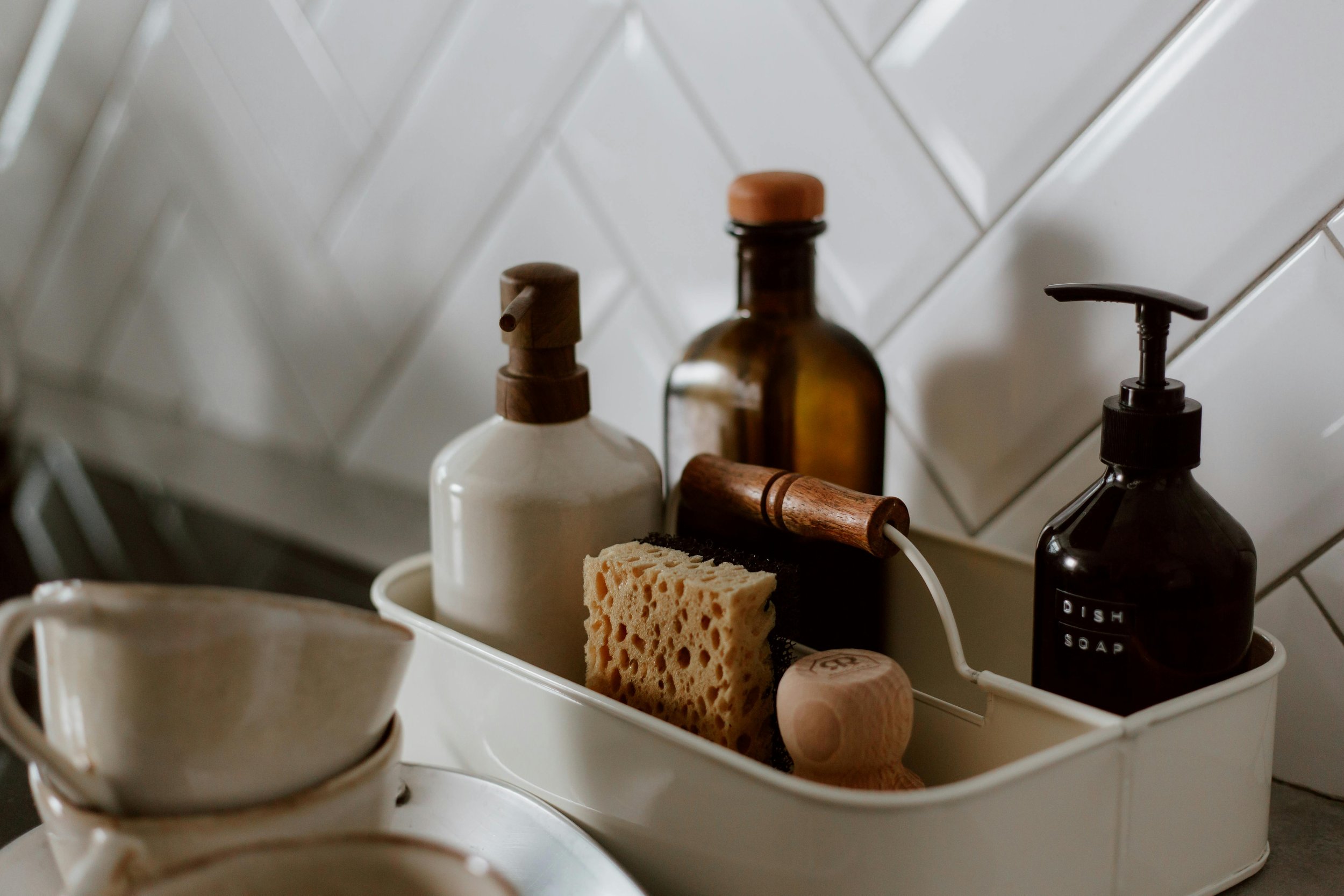 A white container holding various kitchen soap dispensers, a sponge, and a wooden brush, positioned on a countertop with a white tiled backsplash.
