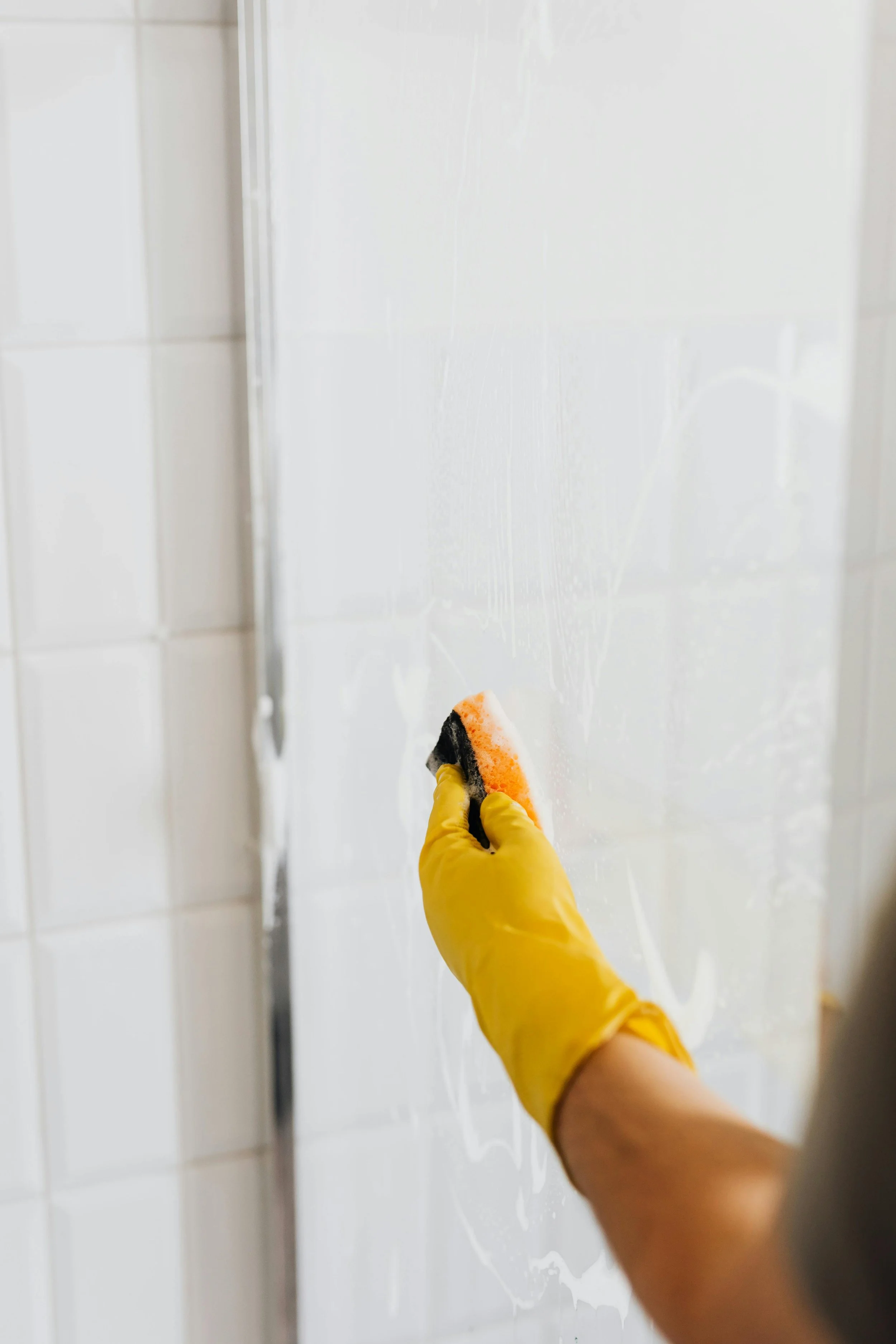 Person wearing a yellow glove cleaning a tiled wall with a sponge and soap.