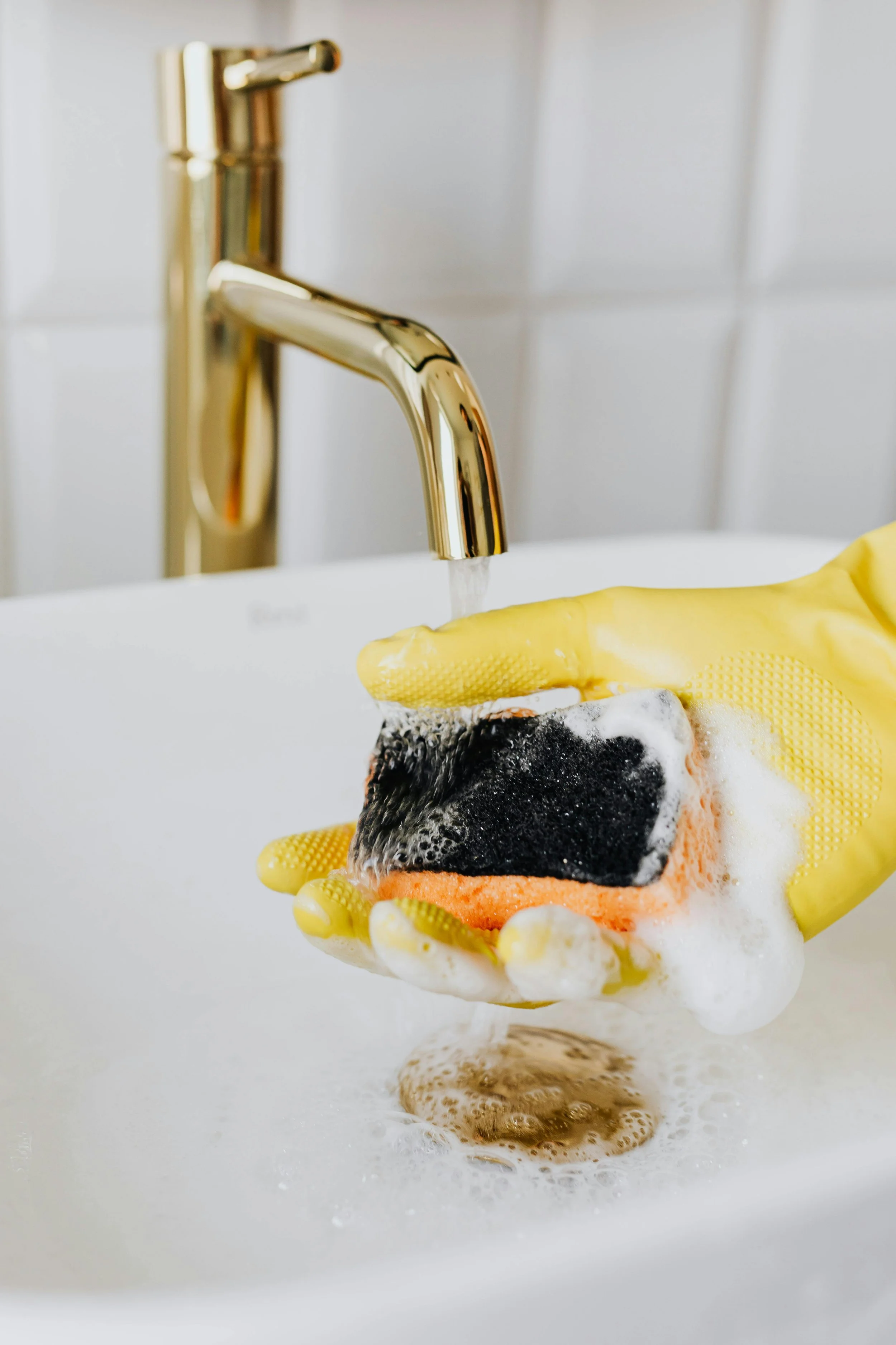 Person wearing yellow rubber gloves washing a black sponge with soap and bubbles in a stainless steel sink