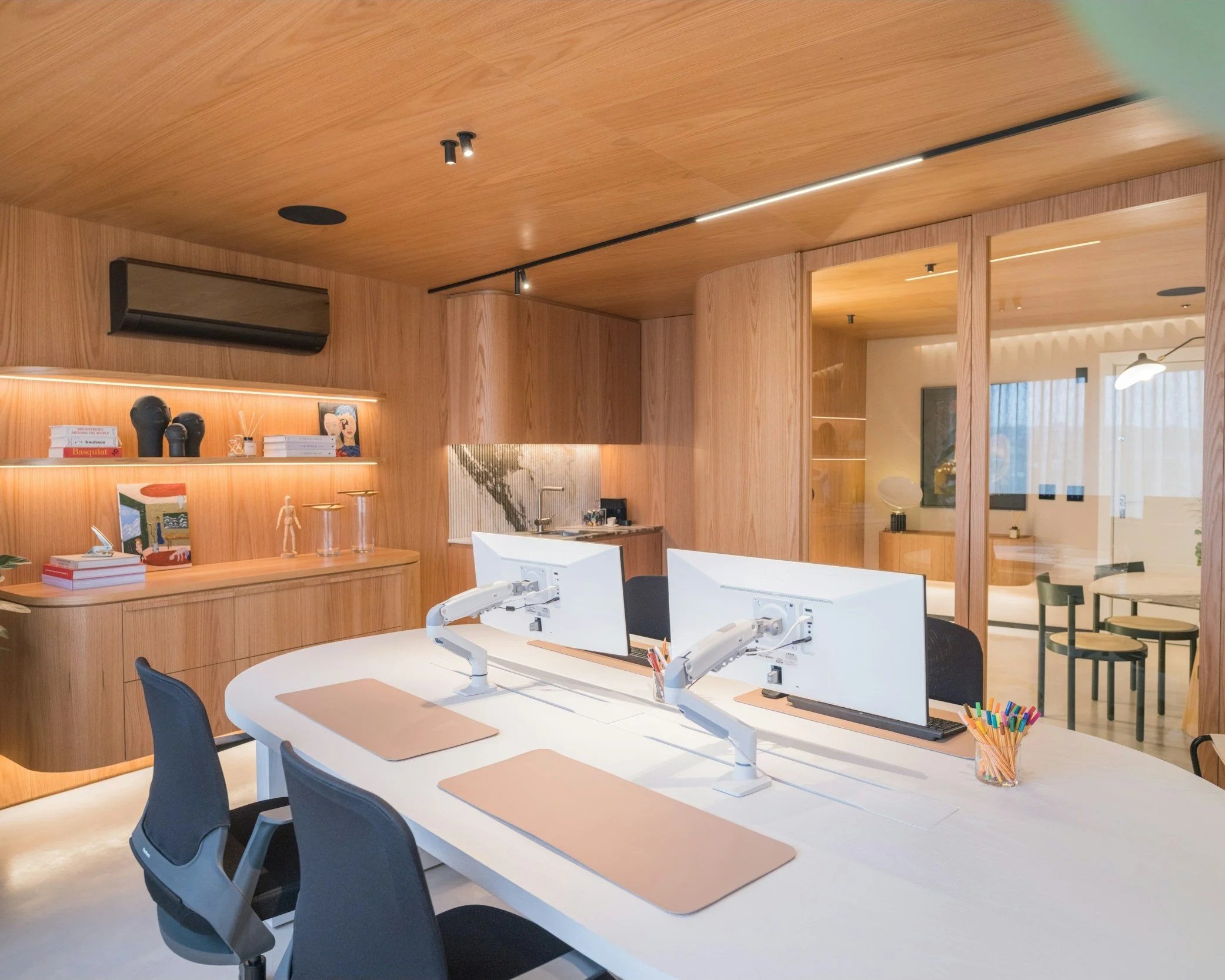 Modern office space with wood-paneled walls and ceiling, featuring a white conference table with four black chairs, dual computer monitors, and a clear cup filled with colored pens. Shelves on the wall display books and decorative items, and a glass partition separates a small lounge area with additional furniture.