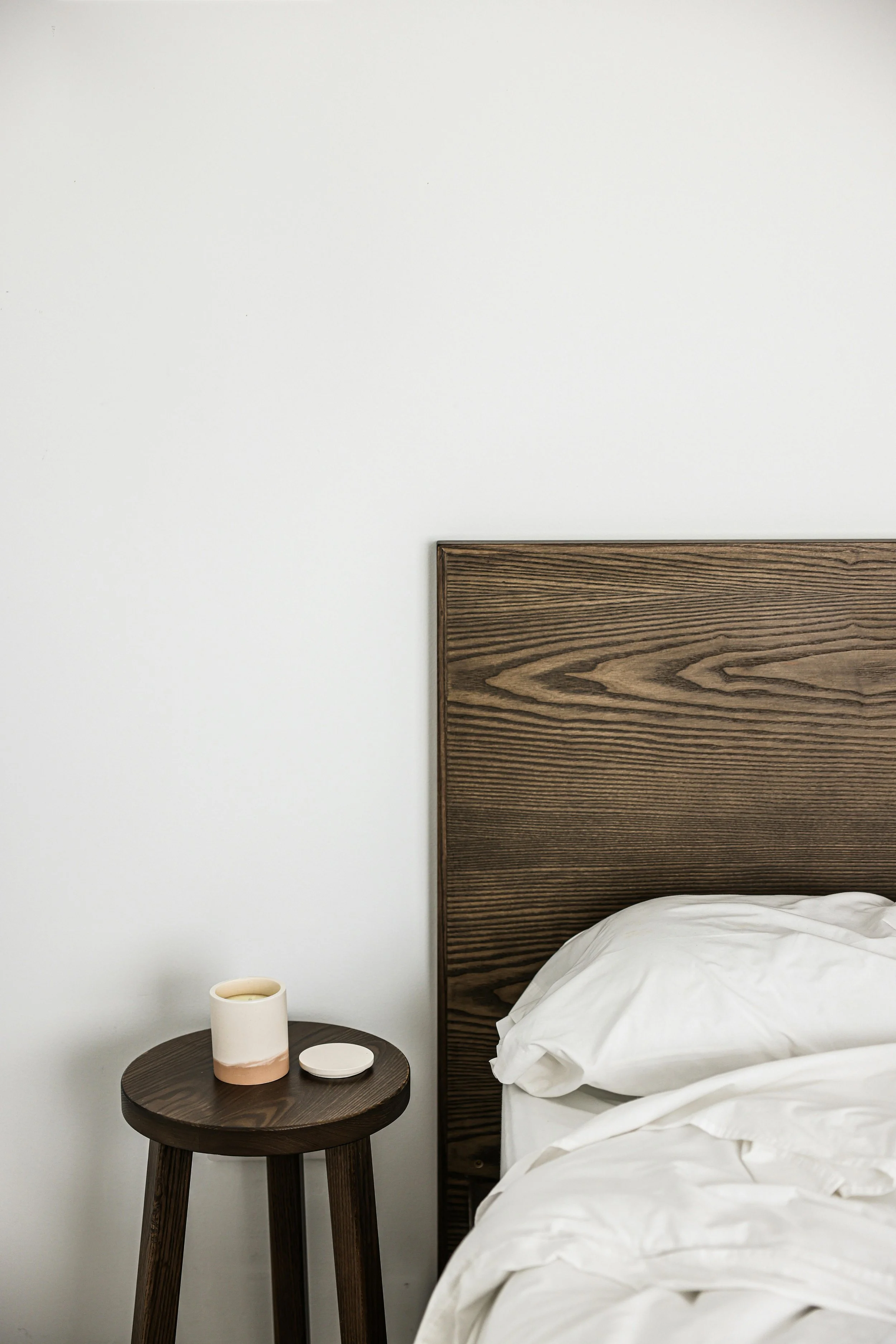 A bedroom scene with a white wall, a wooden headboard, a bed with white bedding, and a small dark wooden bedside table holding a beige ceramic mug and a small round object.