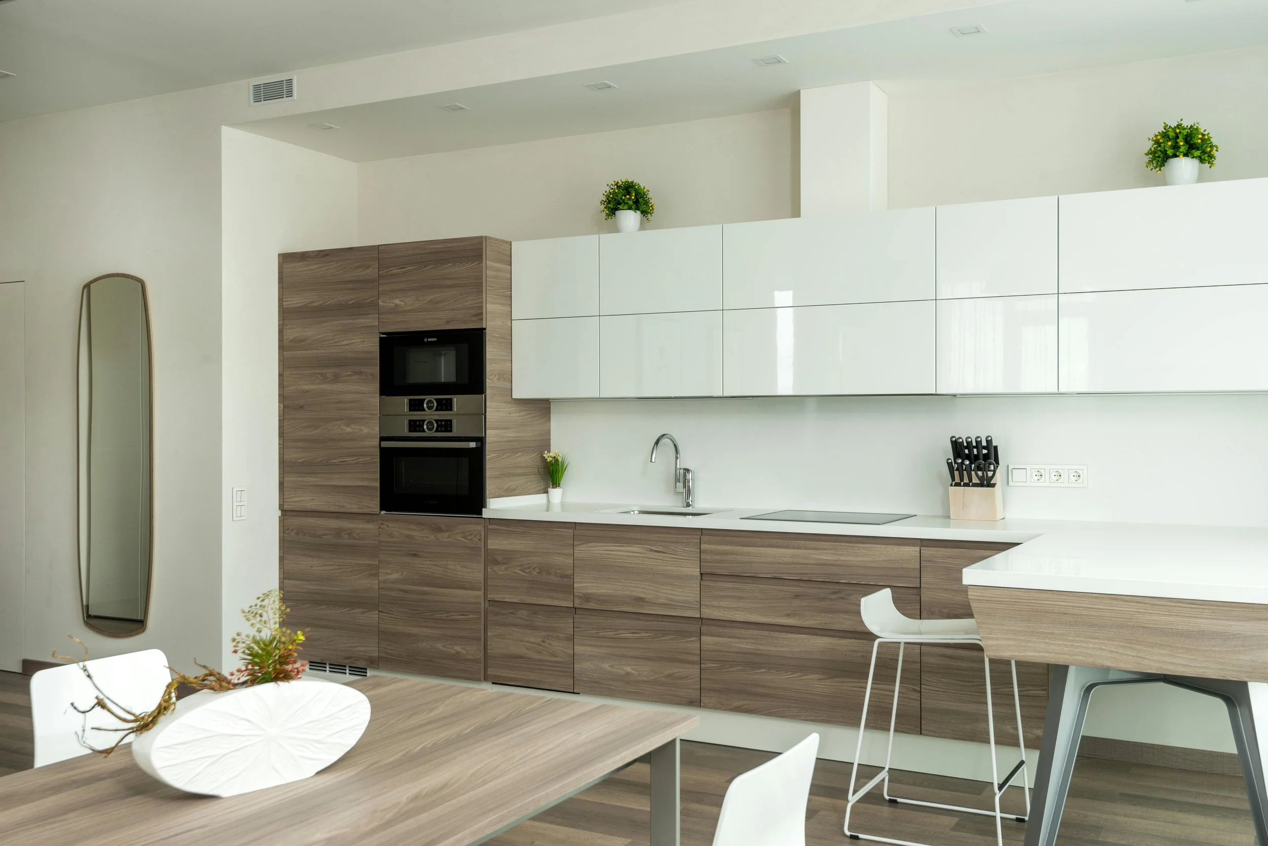 Modern kitchen with wood and white cabinetry, integrated appliances, a white countertop with a sink, a knife block, green potted plants, and a wooden dining table with white chairs.