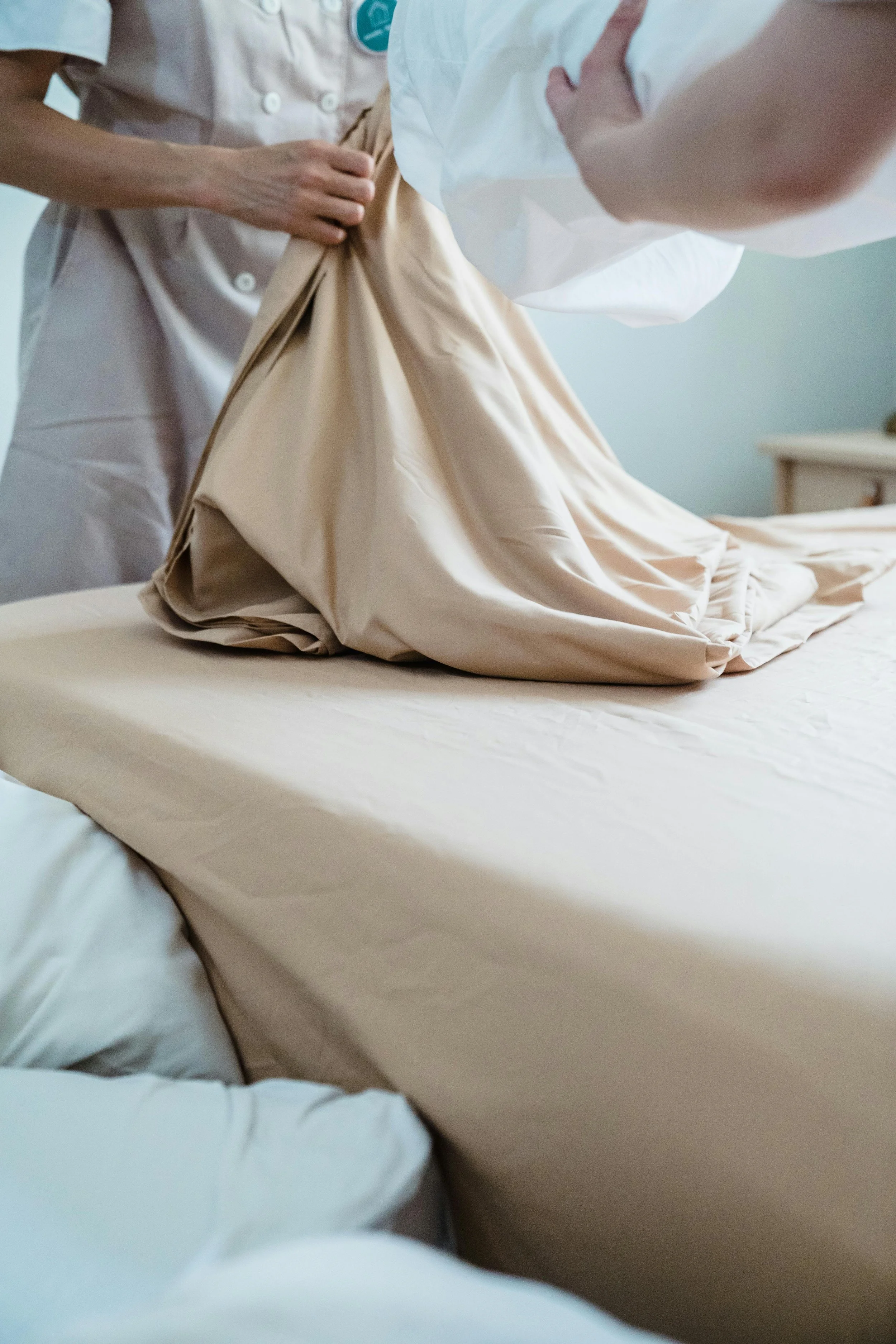 Two healthcare workers making a bed with beige sheets in a hospital or medical facility.
