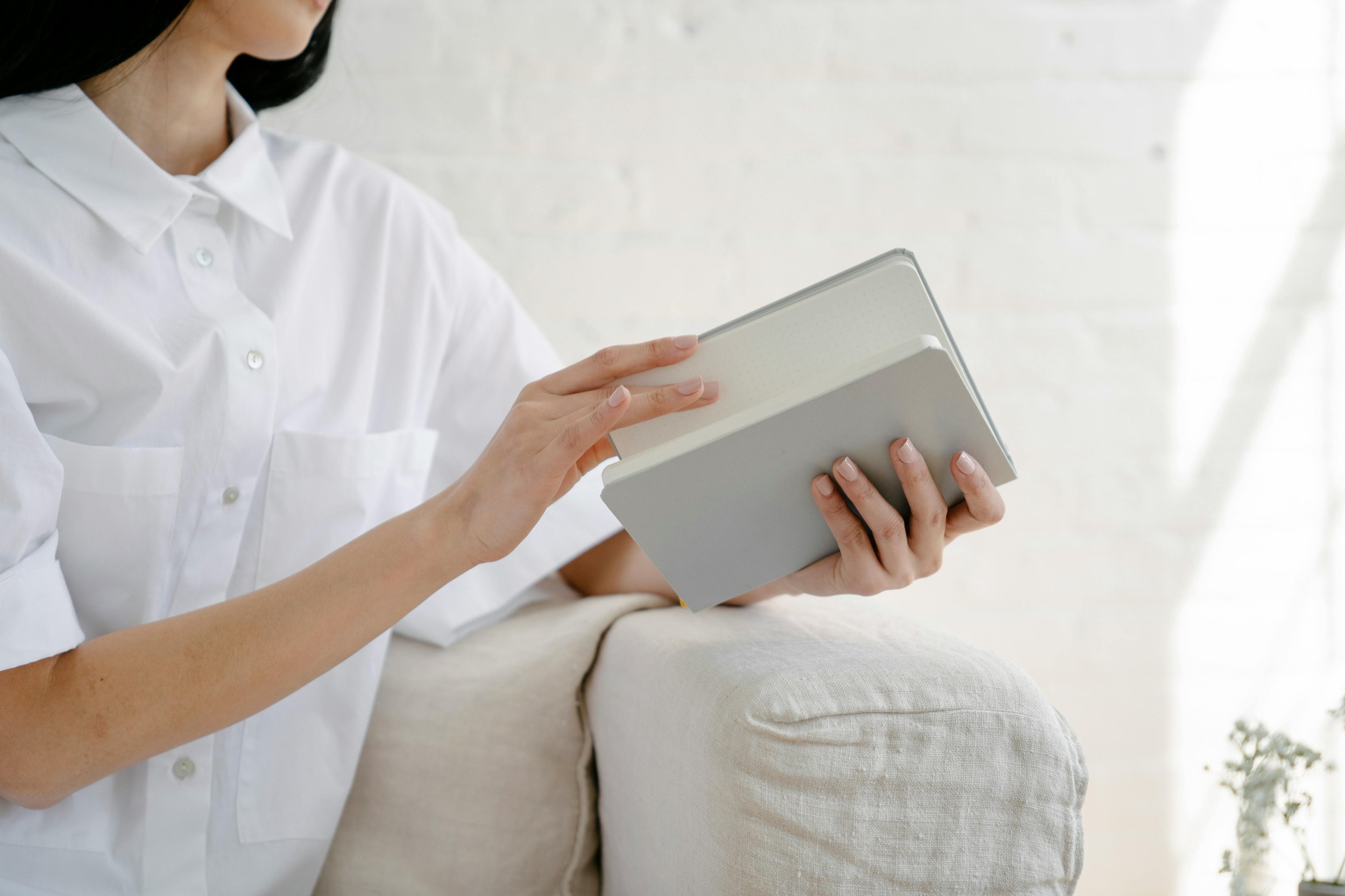 A woman in a white shirt holding a tablet and a closed notebook, standing next to a beige sofa in a bright room.