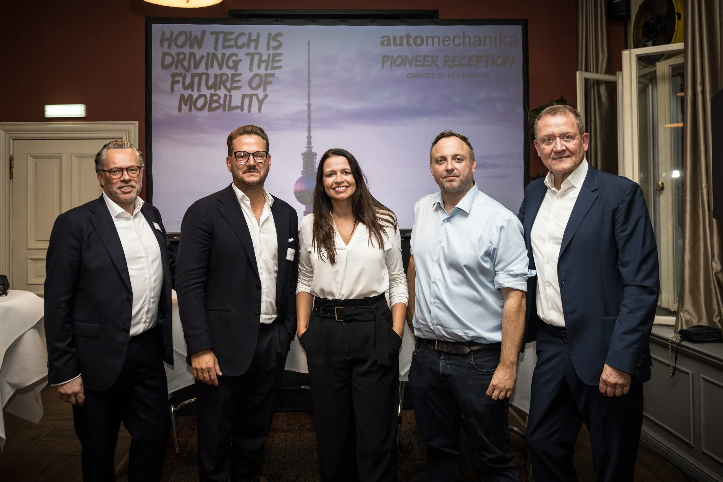Six people in business attire standing together indoors in front of a presentation screen that reads 'How Tech is Driving the Future of Mobility' and 'automechanika Pioneer Reception.'