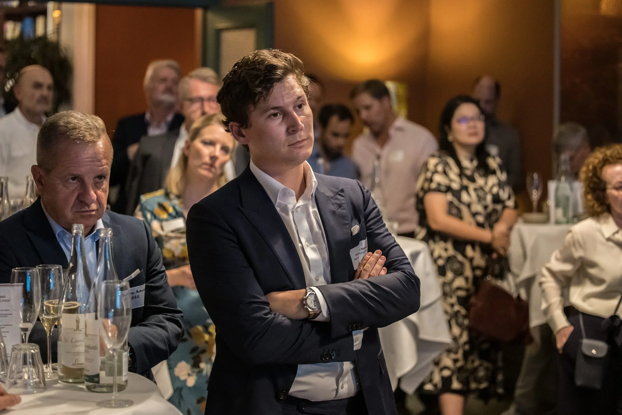 A group of people attending a formal event or conference, with a young man in a navy suit standing with arms crossed, listening attentively.