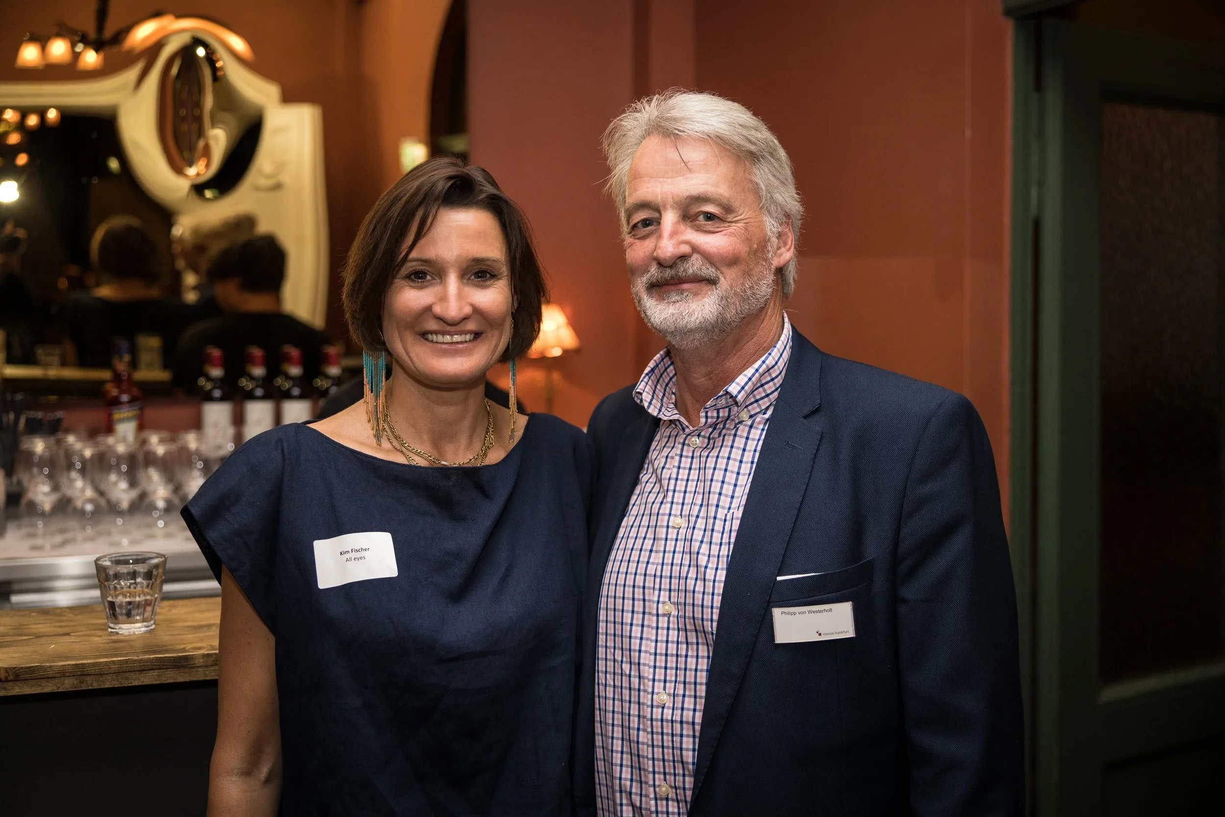 A woman and a man smiling at a social event, standing indoors near a bar with bottles and glasses in the background, both wearing name tags.