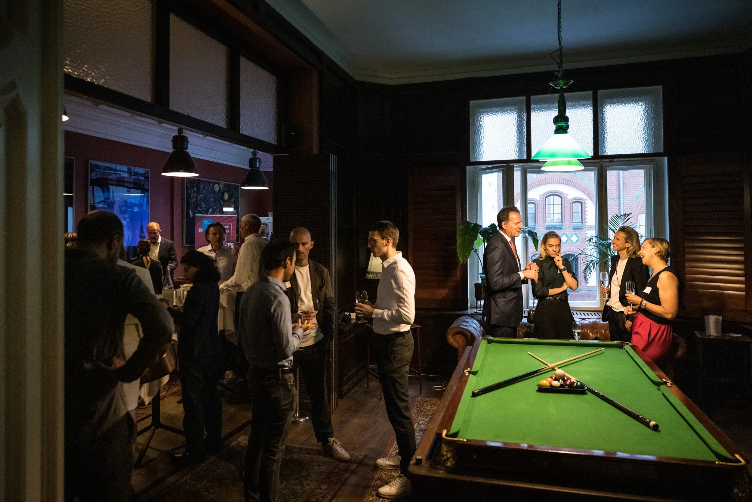 People socializing in a dimly lit room with a pool table near large windows, some holding drinks, with artwork on the walls and a vintage ambiance.