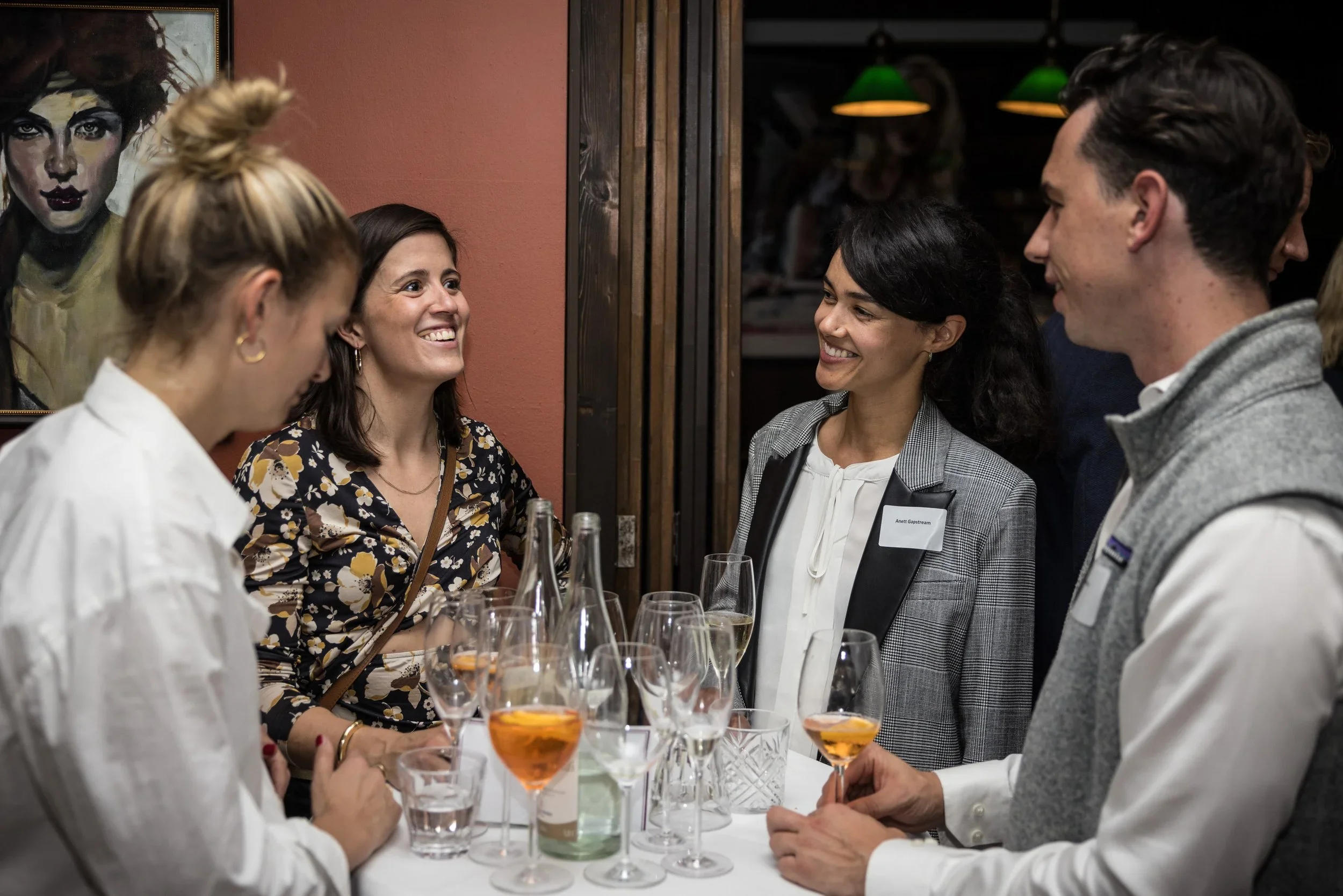 Group of five people having a conversation at a social gathering with drinks on a table, including wine glasses and cocktails.