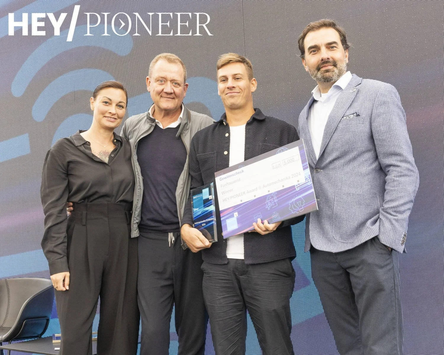 A group of four people standing in front of a screen that says 'HEY PIONEER' with Wi-Fi symbols, with one person holding a large check or award certificate.