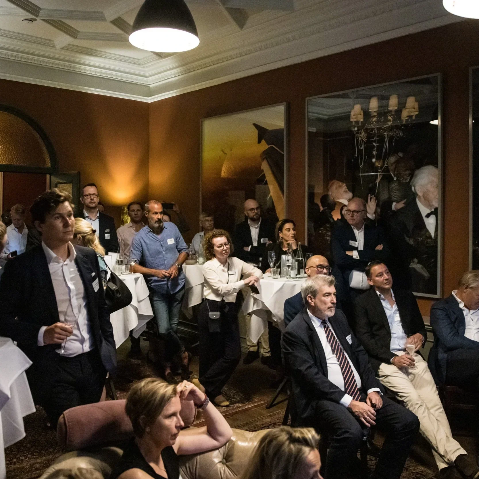 Group of people attending a formal event, standing and sitting in a dimly lit room with brown walls, large paintings, and ornate ceiling details.