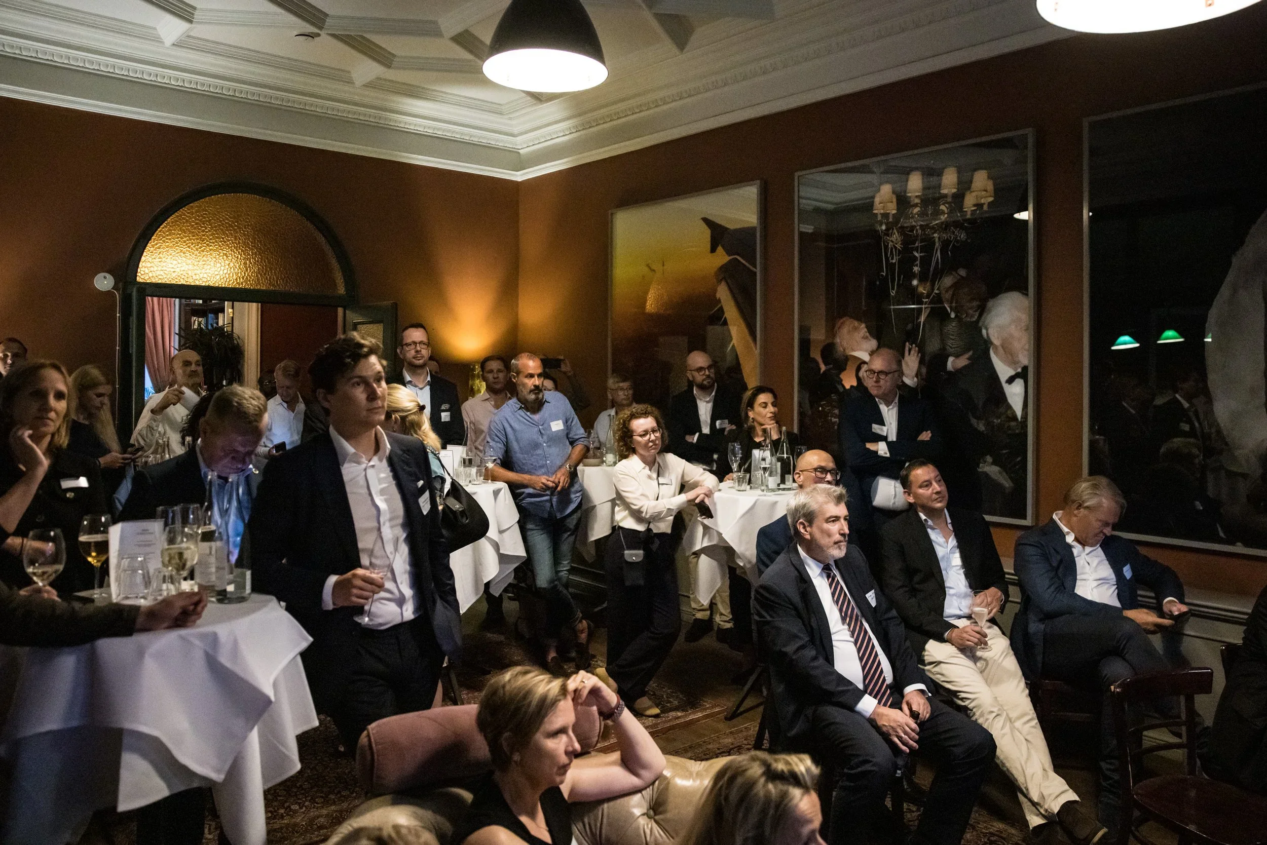 People attending a formal event in a dimly lit room with brown walls, large paintings, and tables with white tablecloths, glasses, and drinks.