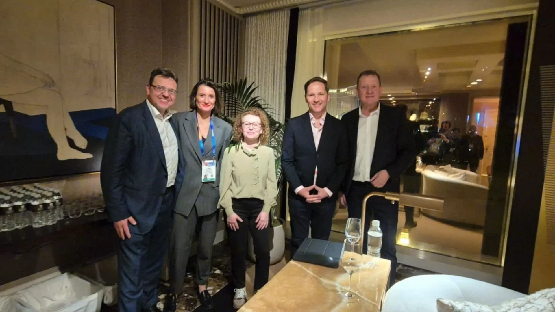 A group of five professionally dressed people standing together in a hotel or conference room, smiling at the camera.