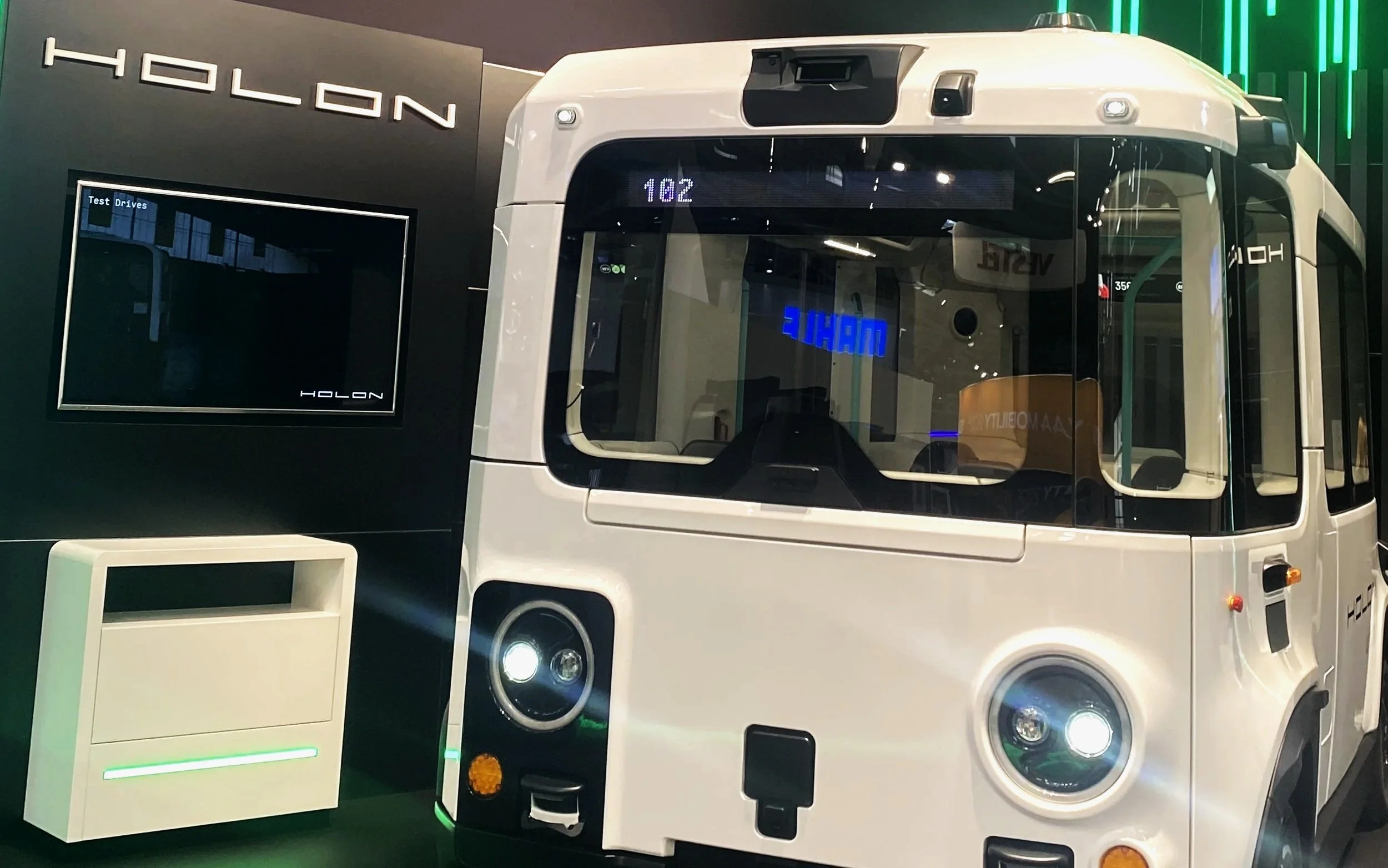 A white autonomous shuttle bus with black accents on display at a technology exhibit, with a black wall behind it displaying the brand name 'Holon' and a monitor showing the company's logo.
